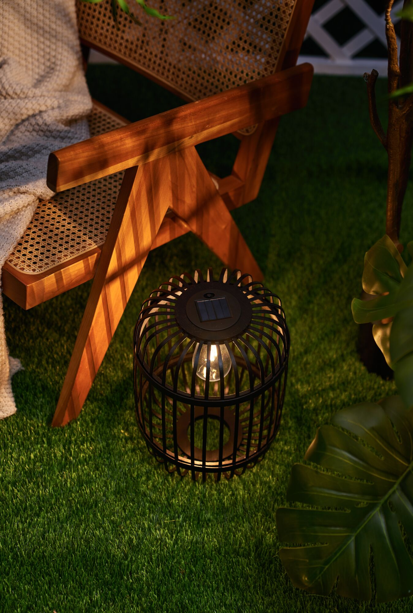 Solarna latarnia ogrodowa na sztucznej trawie obok drewnianego krzesła.