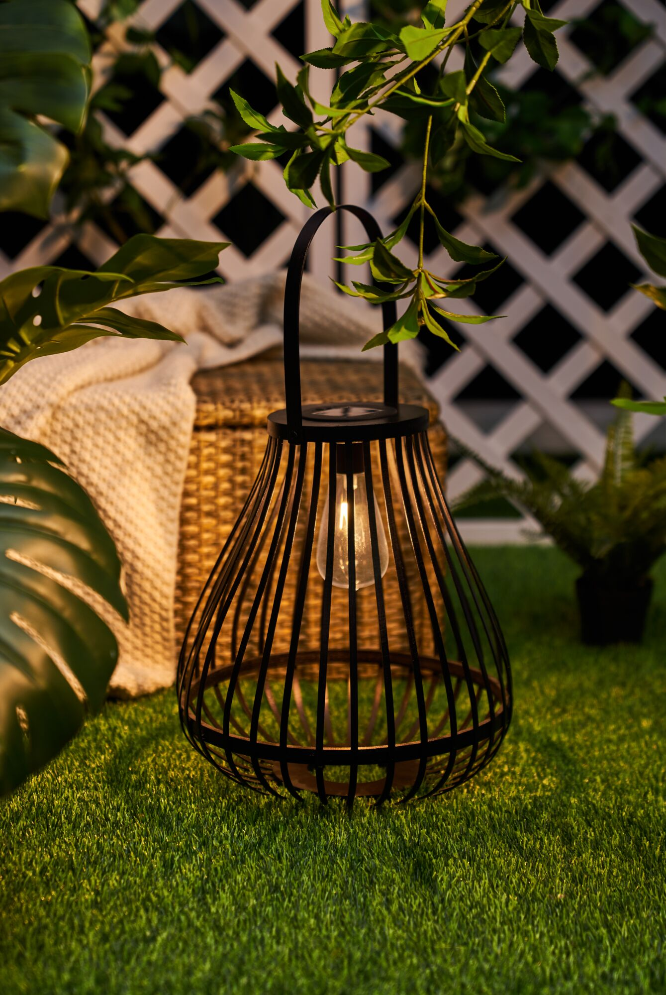 Czarna latarnia z żarówką stoi na sztucznej trawie obok kosza i koca.