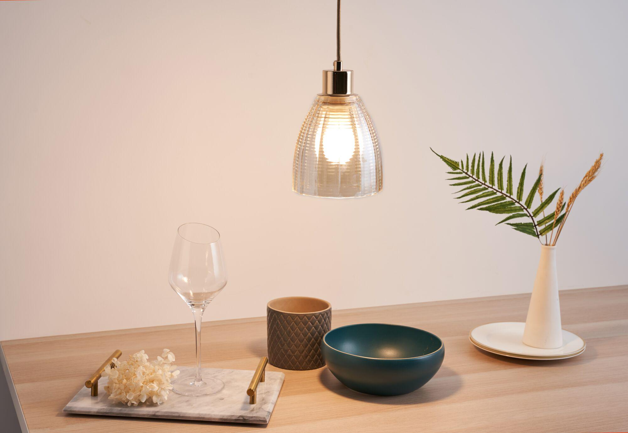 Lampa wisząca oświetla kompozycję ze szkła, miski, wazonu i rośliny.