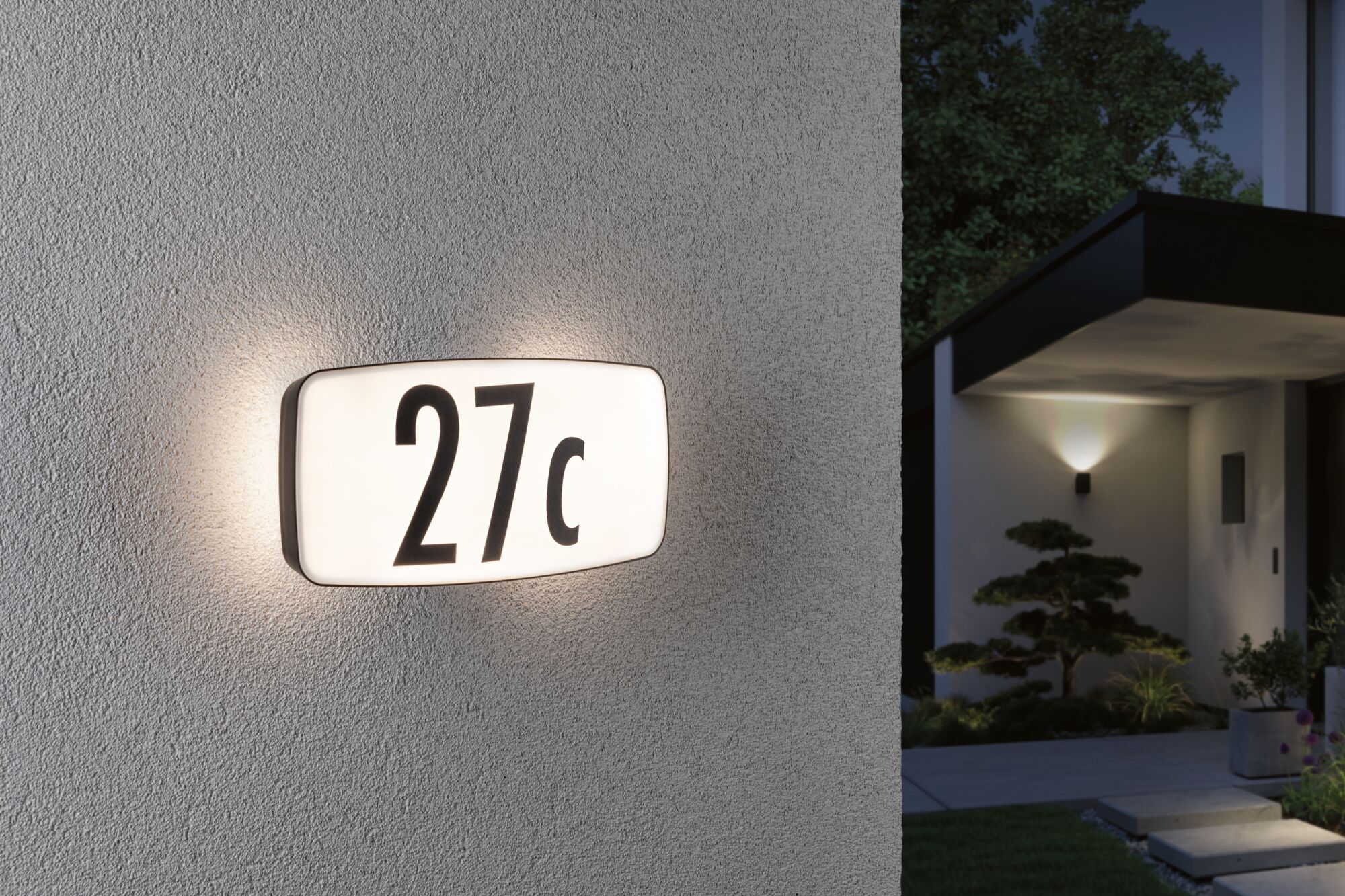 Beleuchtetes Hausnummernschild mit 27c an einer grauen Wand, daneben ein Haus.