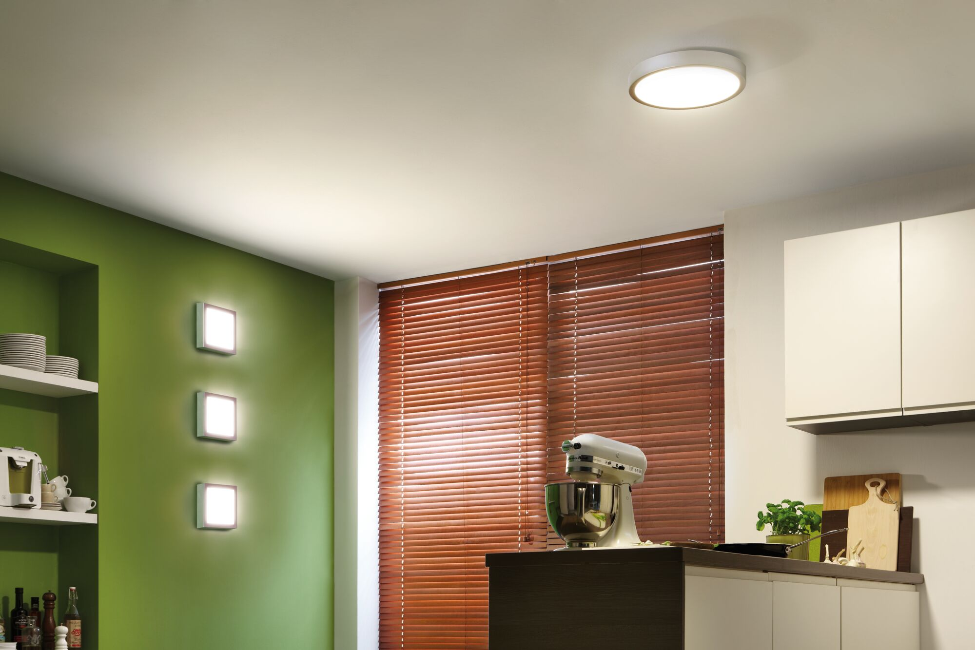 Cucina moderna con parete verde, tende alla veneziana, impastatrice e mobili bianchi.