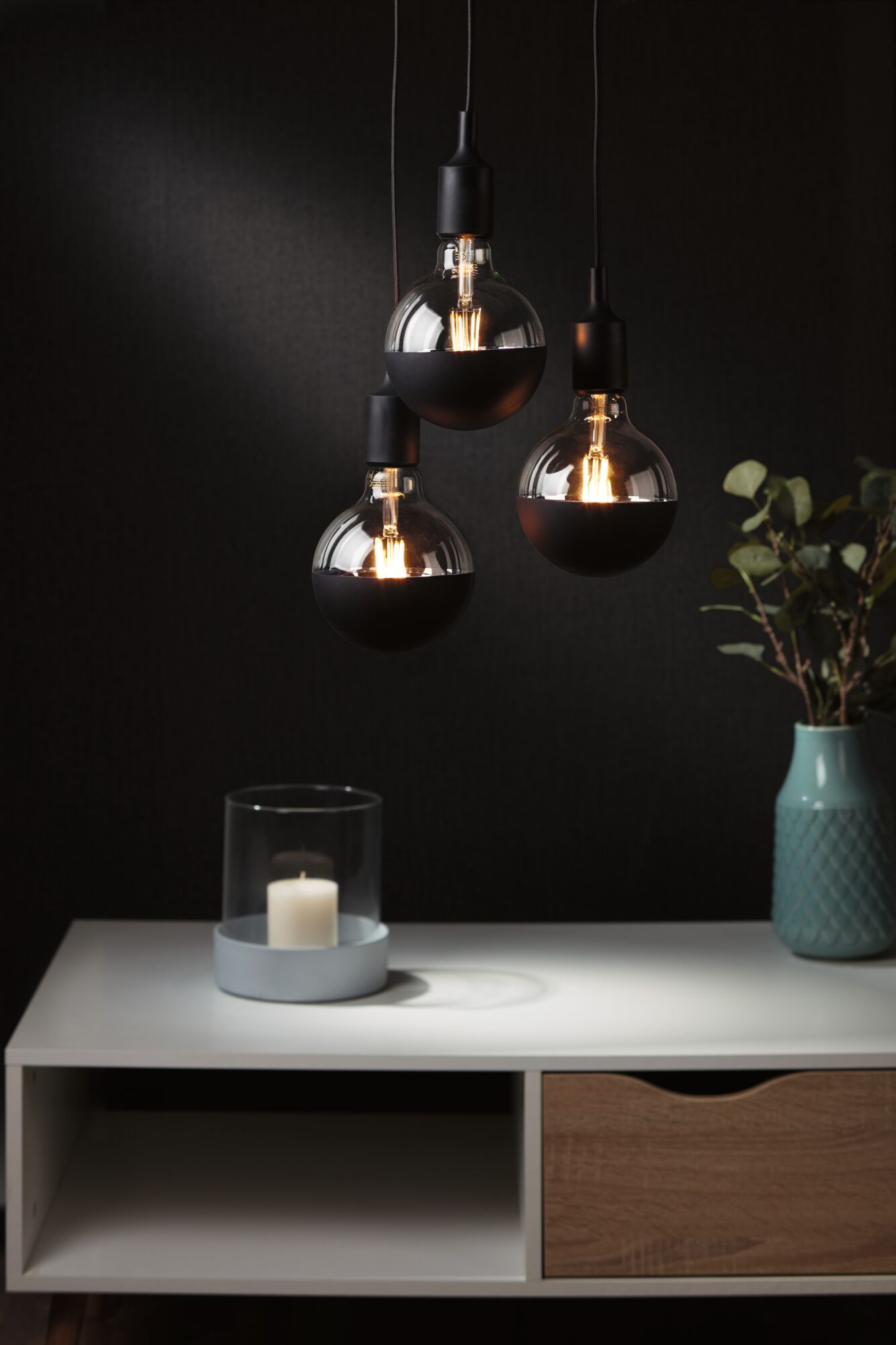 Trzy lampy wiszące z czarnymi kapturkami i szklanymi żarówkami nad stołem ze świecą i wazonem.