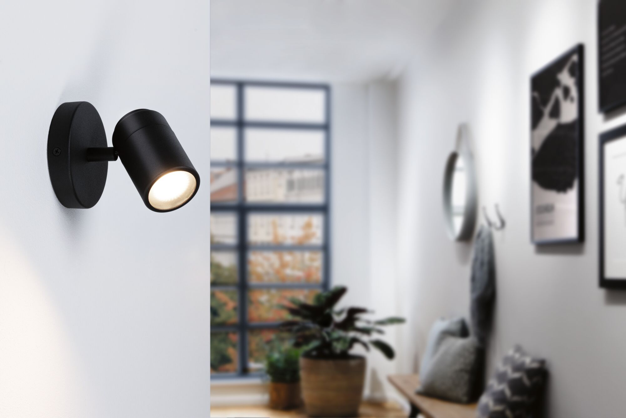 Czarny kinkiet świeci na białej ścianie; korytarz z roślinami i obrazami.
