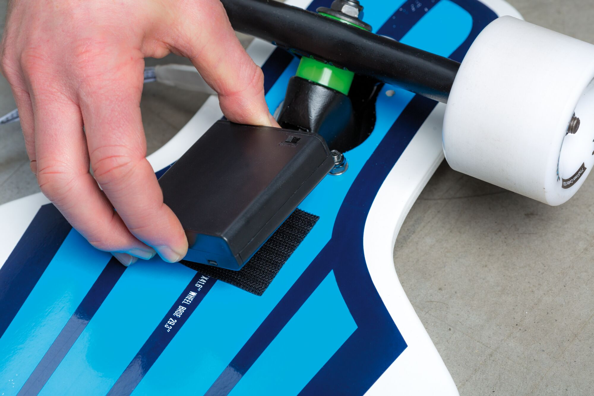 Hand drückt schwarzes Kästchen auf Skateboard mit blau-weißer Grafik.