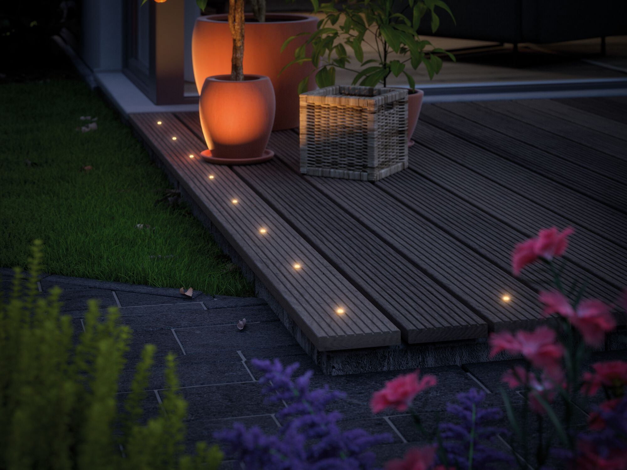 Ciemny drewniany taras z wbudowanymi światłami, roślinami i kwiatami o zmierzchu.