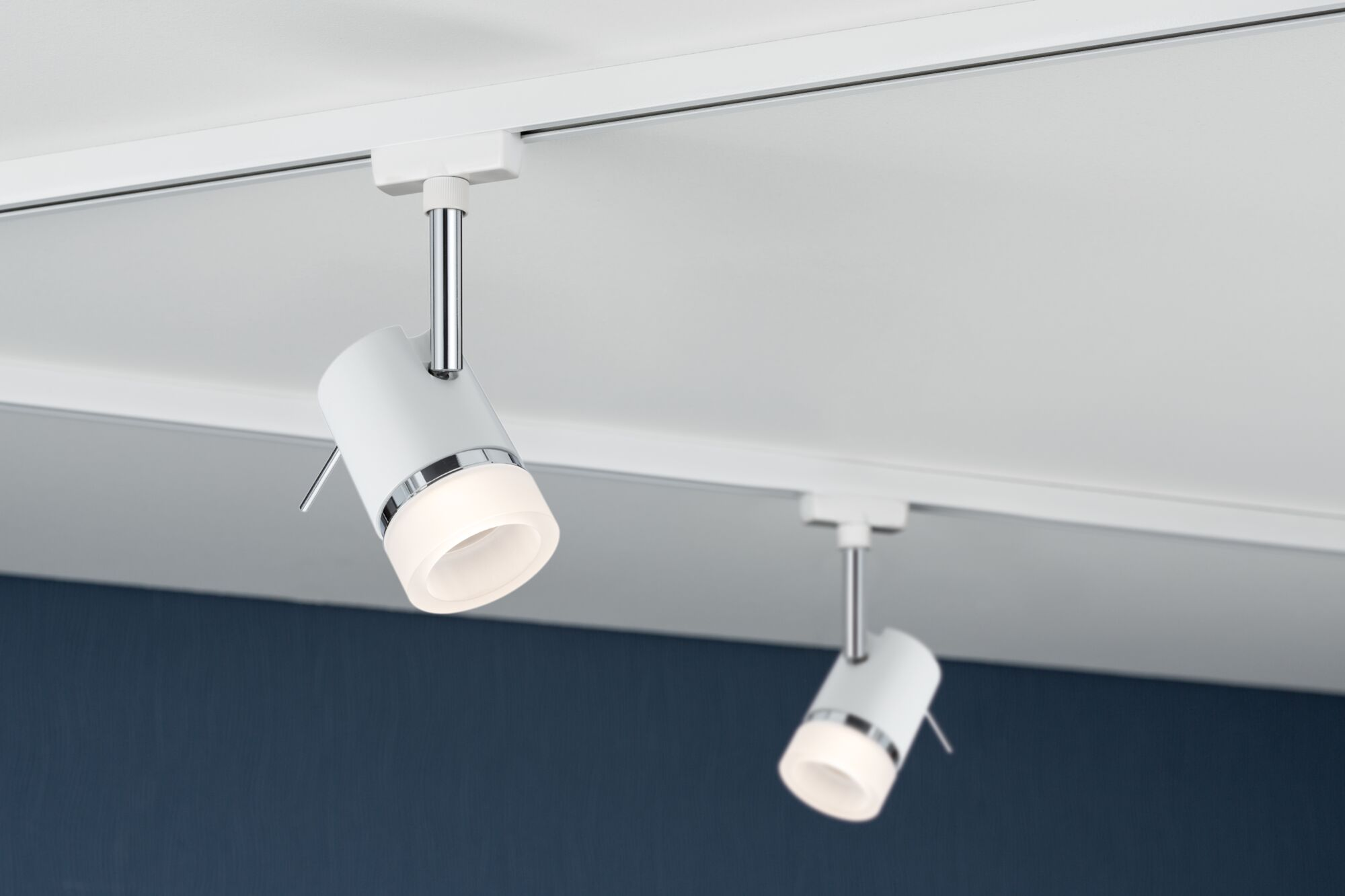Zwei weiße zylindrische Schienenstrahler mit Chromakzenten sind auf einer weißen Schiene vor einer dunkelblauen Wand montiert.