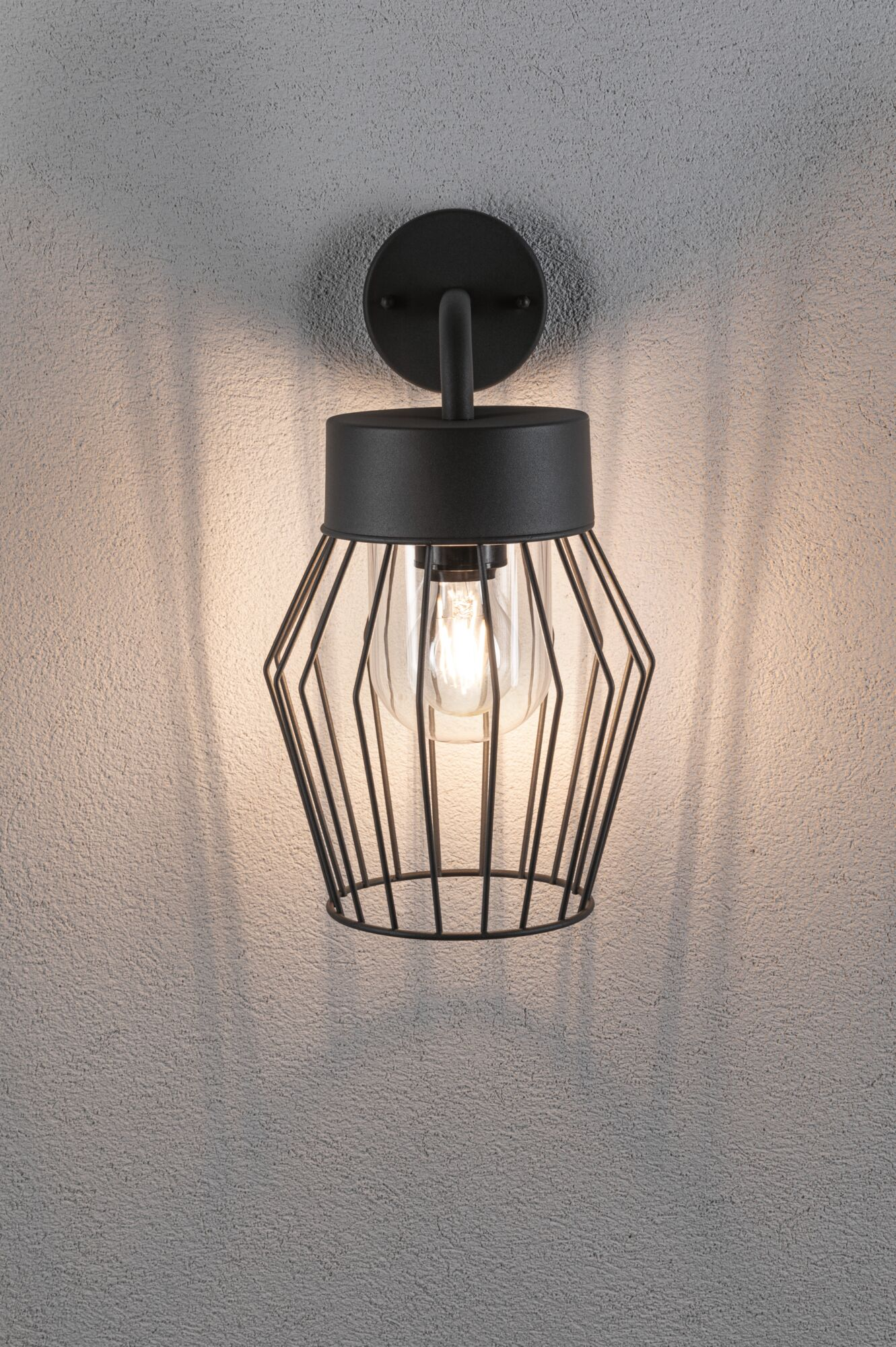 Schwarze Wandleuchte mit Drahtkäfig und Glühbirne an einer strukturierten Wand.