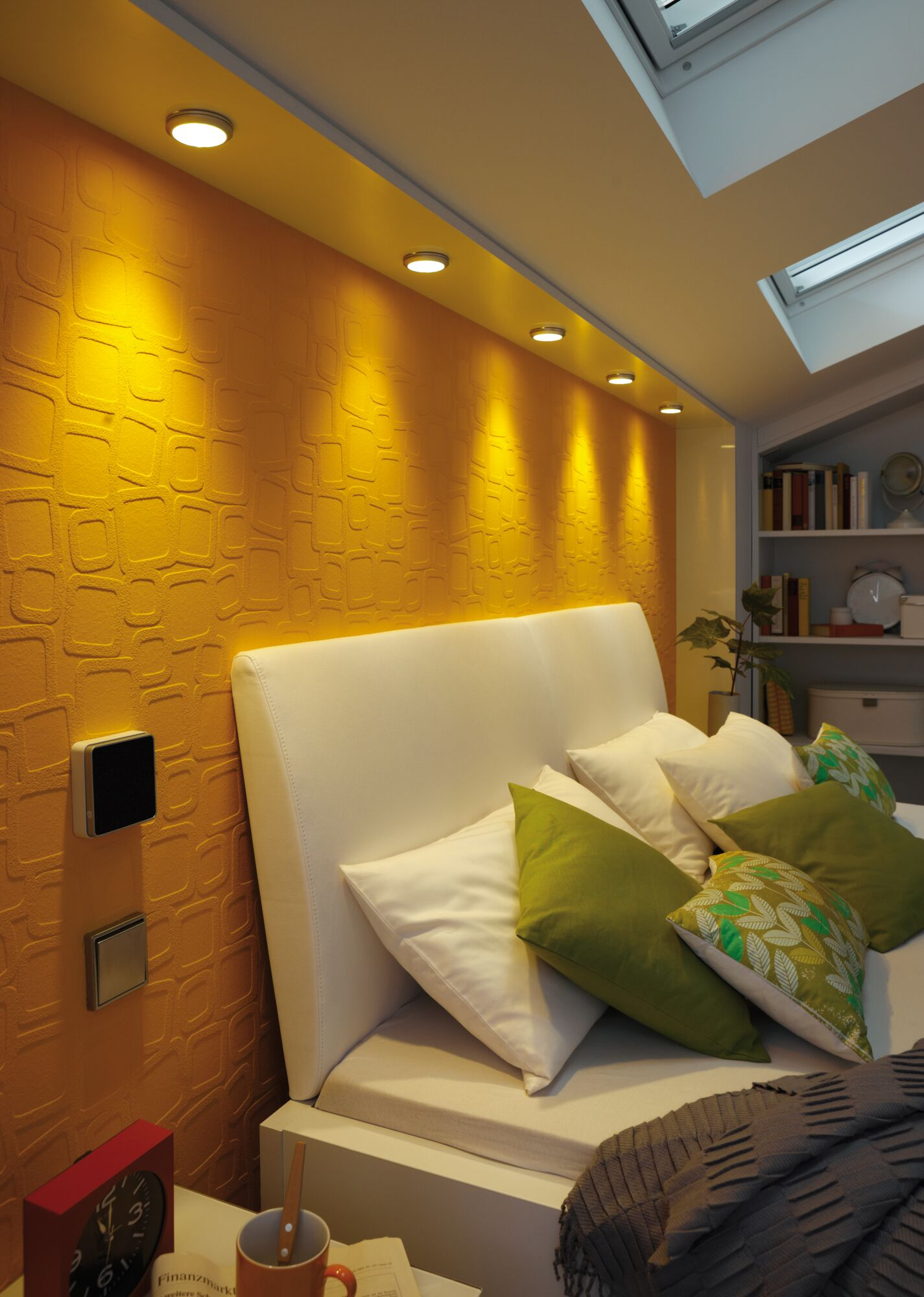 Ein Bett mit weißem Kopfteil und orange gemusterter Wand, beleuchtet von Deckenstrahlern.