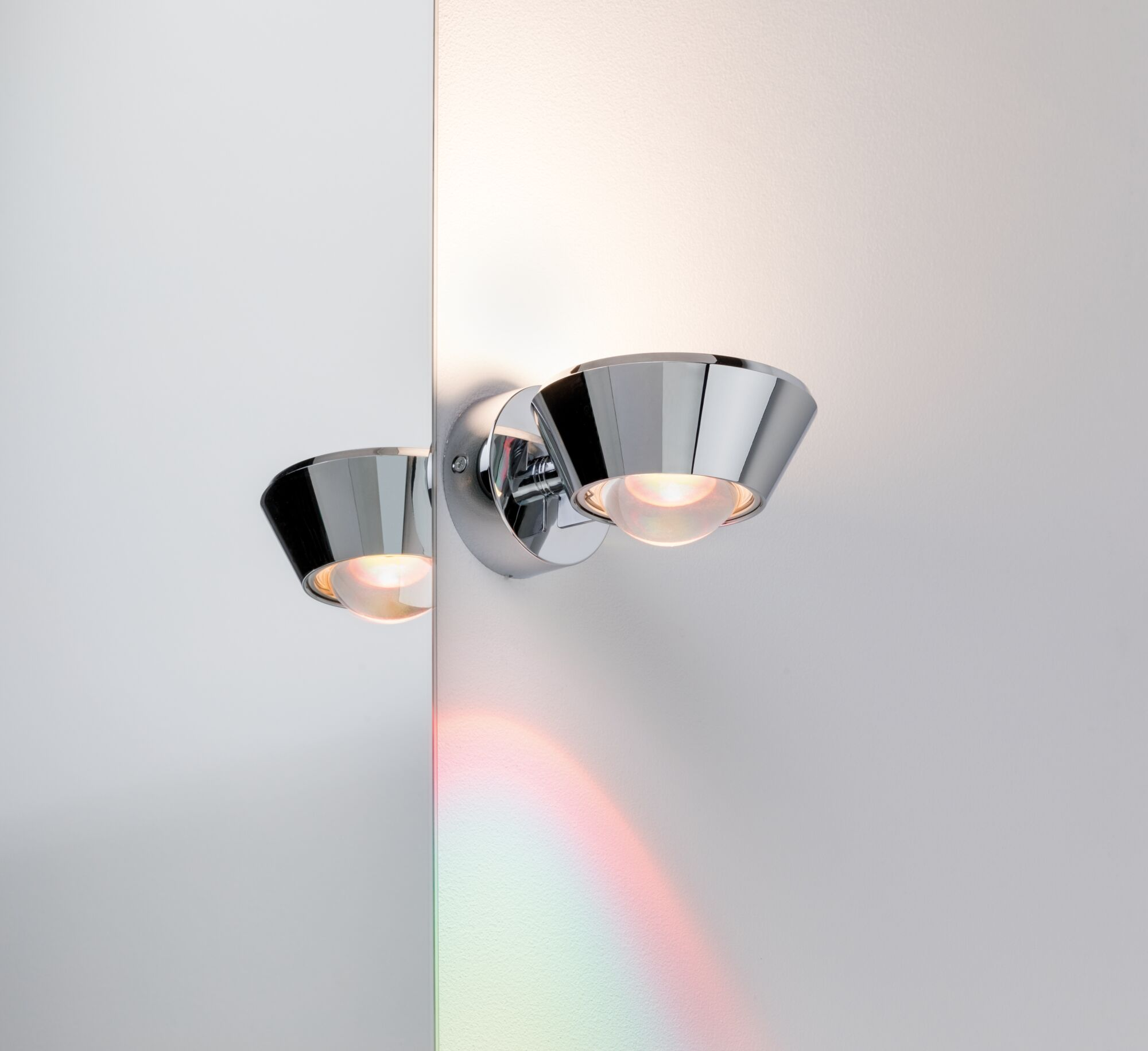 Zwei polierte Chrom-Wandleuchten beleuchten eine weiße Wand mit Regenbogenreflexionen.