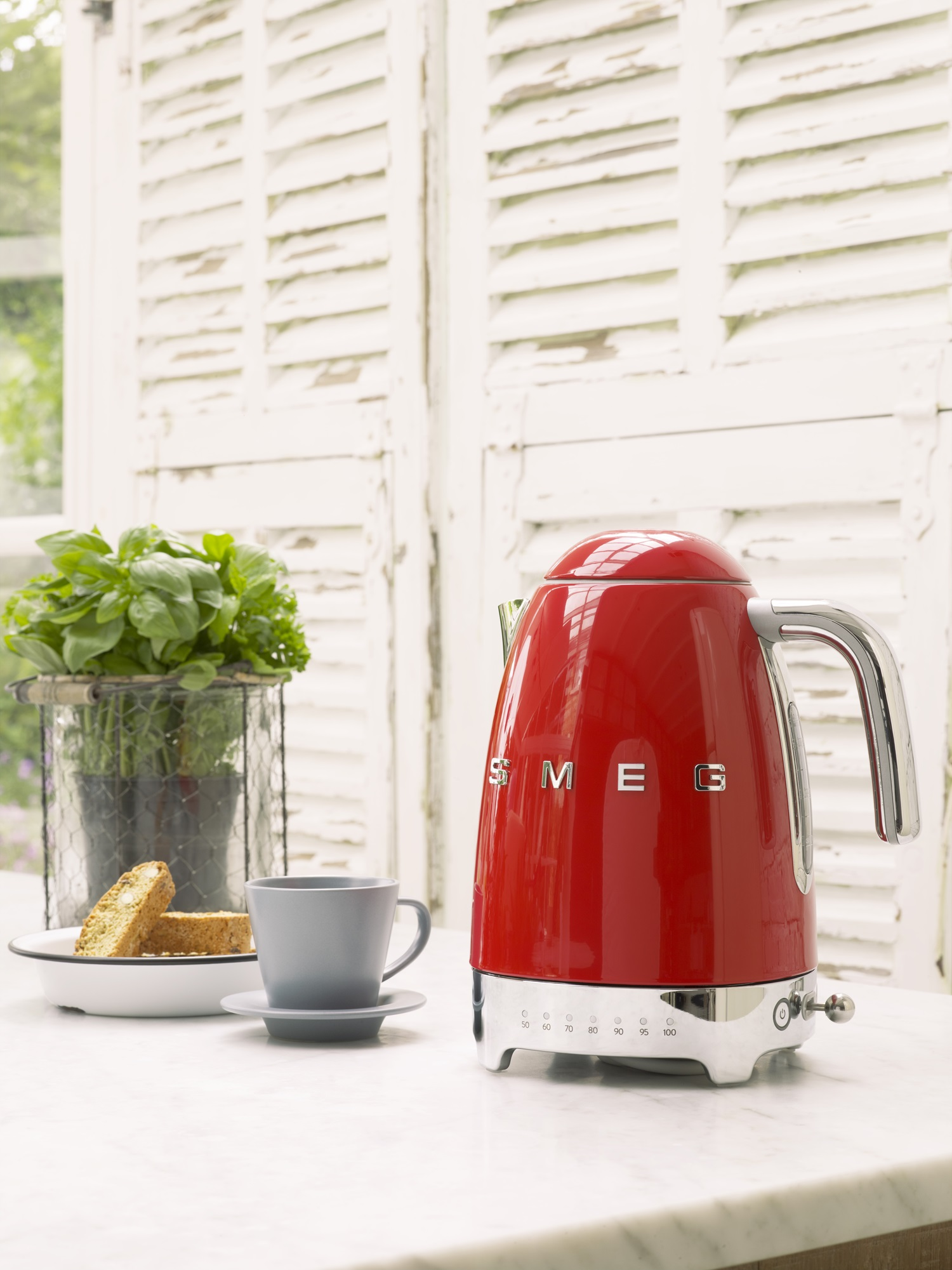Ein roter SMEG-Wasserkocher steht auf einer Marmorplatte neben einer Tasse und Biscotti.