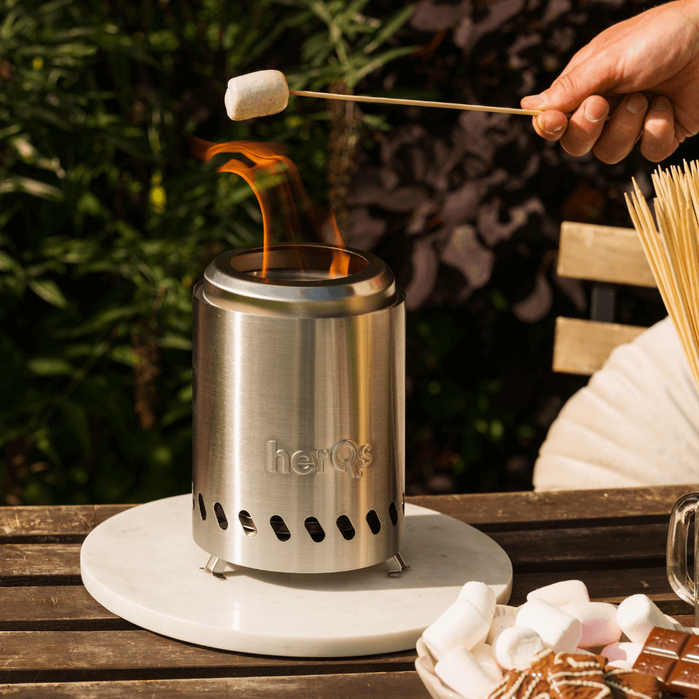 Eine Hand röstet einen Marshmallow über einer kleinen Edelstahl-Feuerstelle mit Flammen.
