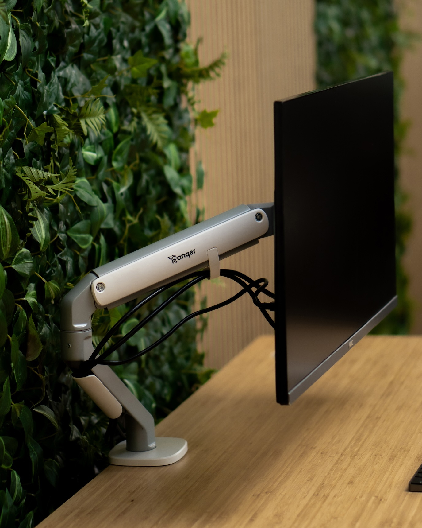 Ein silberner Monitorarm hält einen schwarzen Monitor vor einer grünen Pflanzenwand.