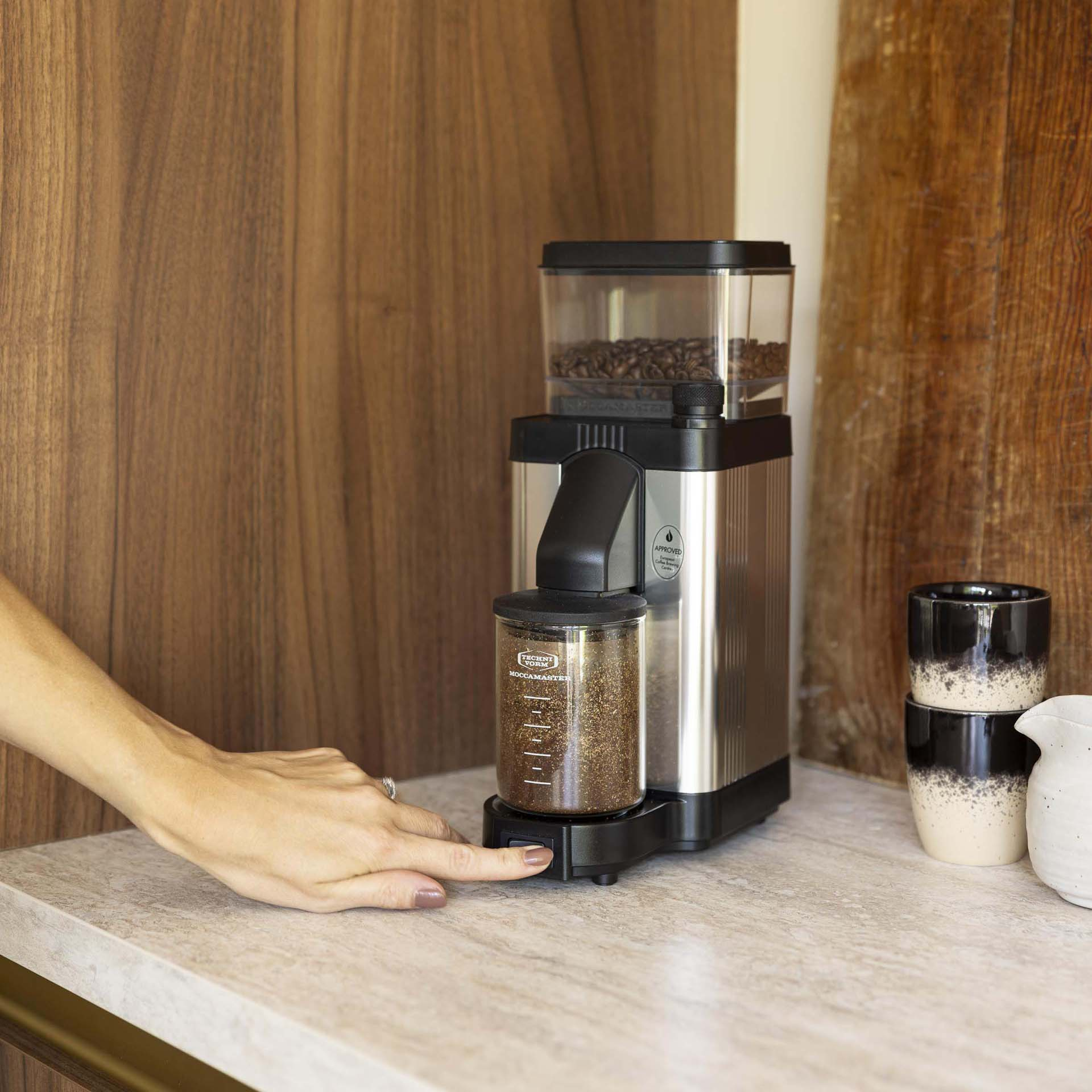 A hand presses a button on a coffee grinder with beans and ground coffee.
