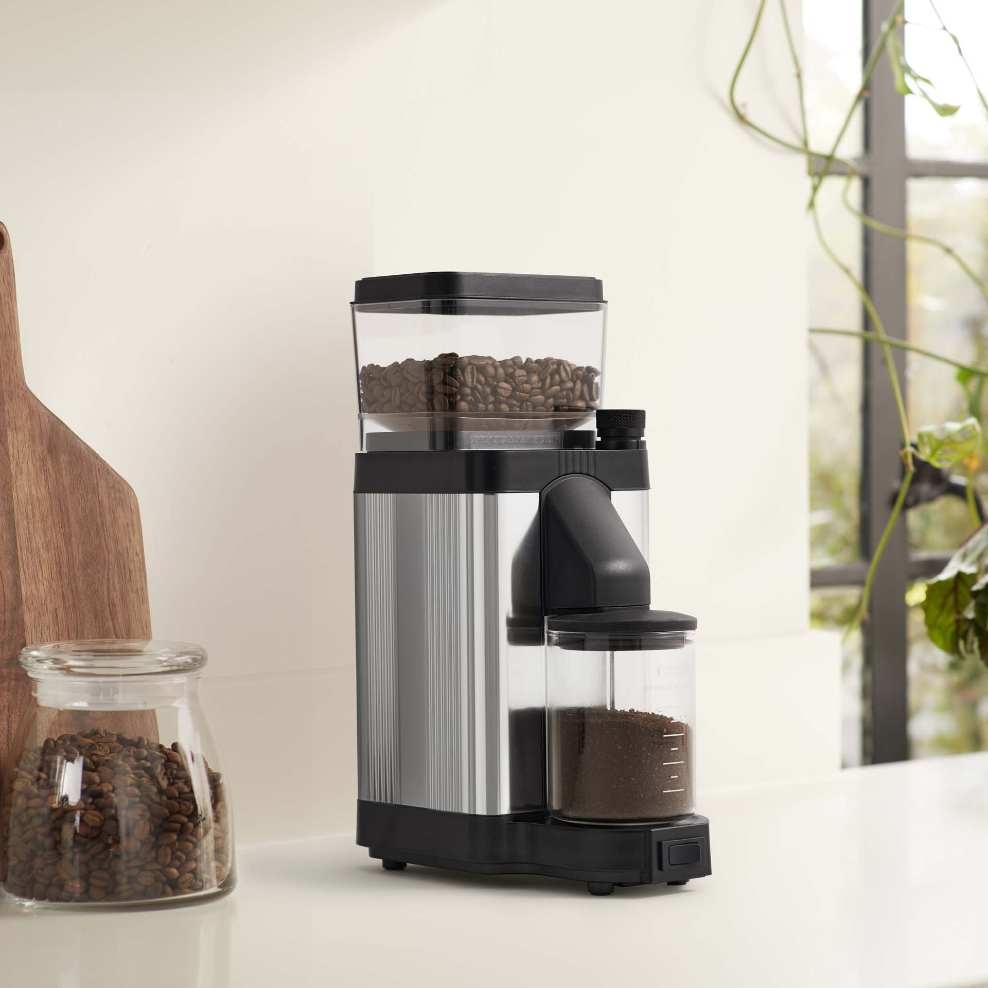 Coffee grinder with beans on top and grounds below, next to a jar of beans.
