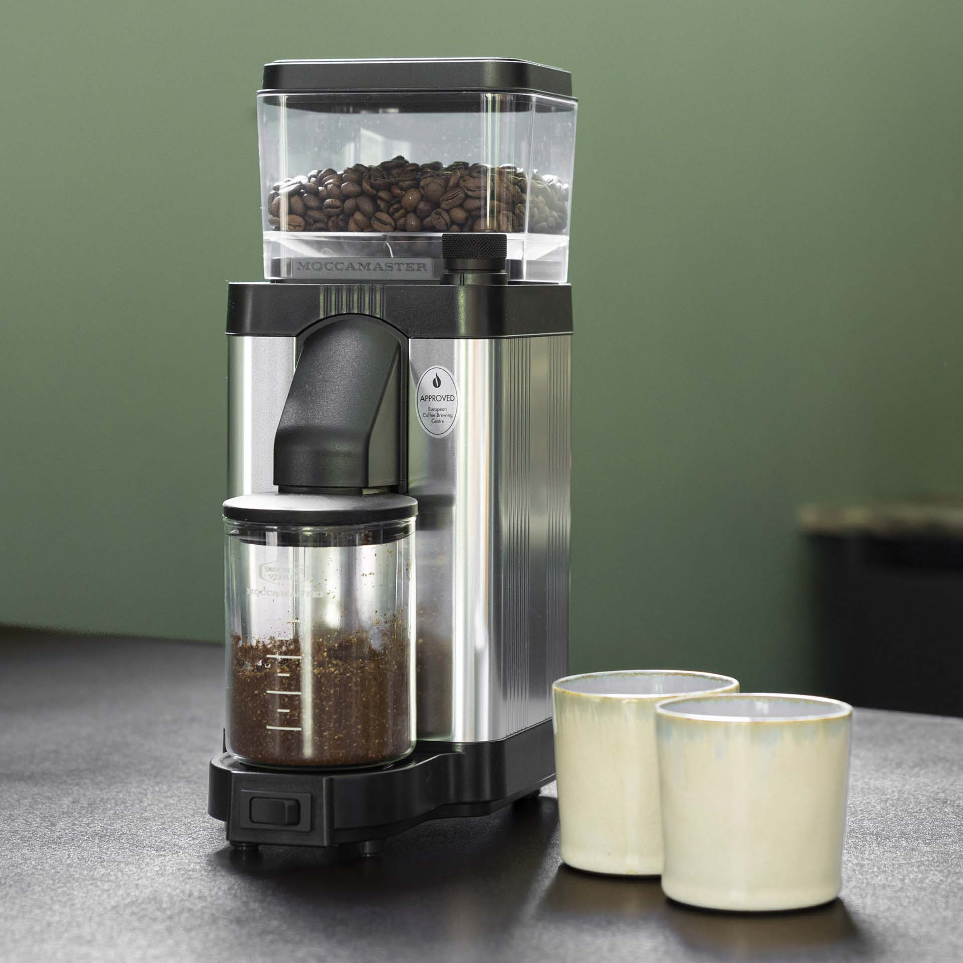 Coffee grinder with beans and ground coffee, with two cups beside it.