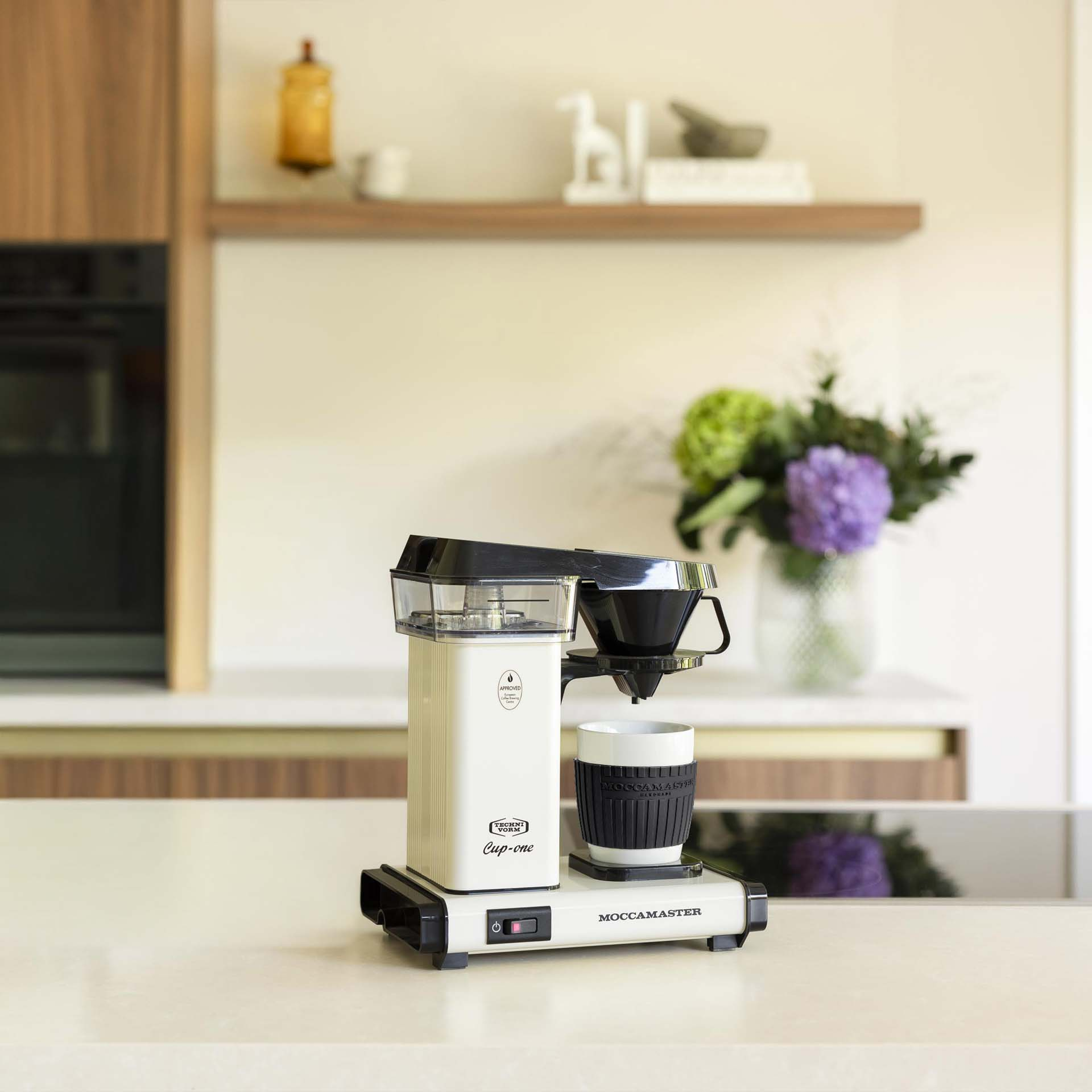 Eine cremefarbene Moccacoffee-Maschine mit schwarzem Filterhalter und Tasse steht auf einer Arbeitsplatte.