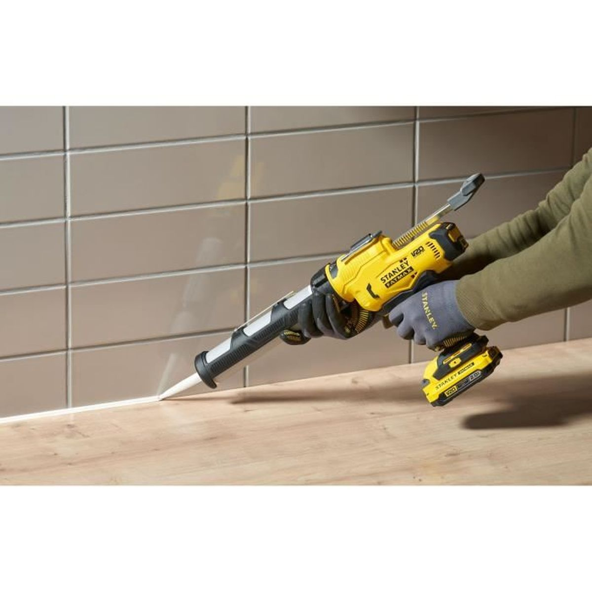 Una mano guantata aziona una pistola per silicone gialla, applicando sigillante bianco su una superficie di legno.