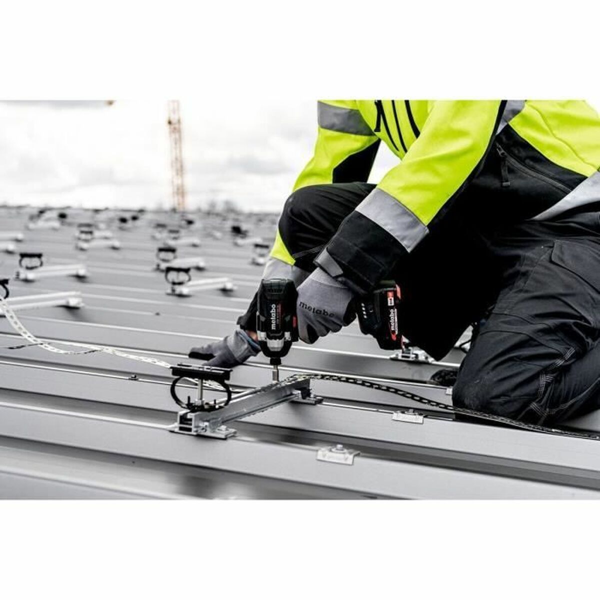 Ein Arbeiter in einer Warnweste installiert mit einem Akkuschrauber Solarpanel-Halterungen auf einem Metalldach.