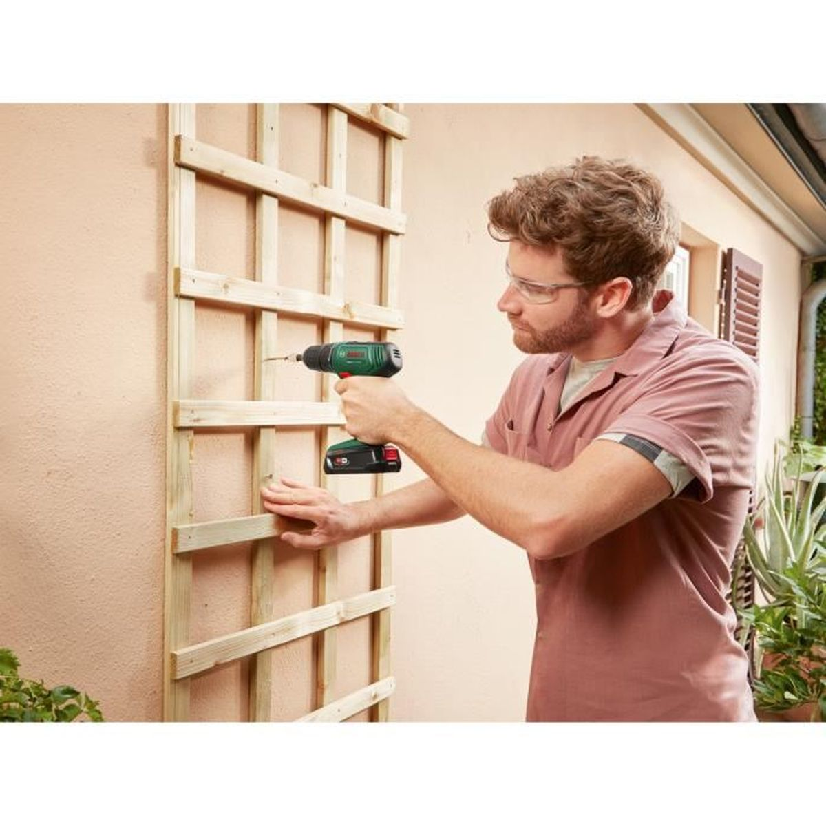 Mężczyzna wierci drewnianą kratkę w ścianie wiertarką.