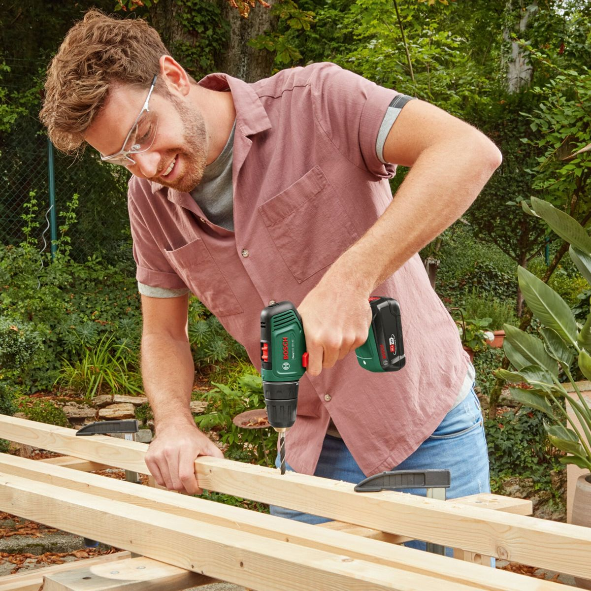 Mężczyzna wierci w drewnie wiertarką Bosch, nosi okulary.