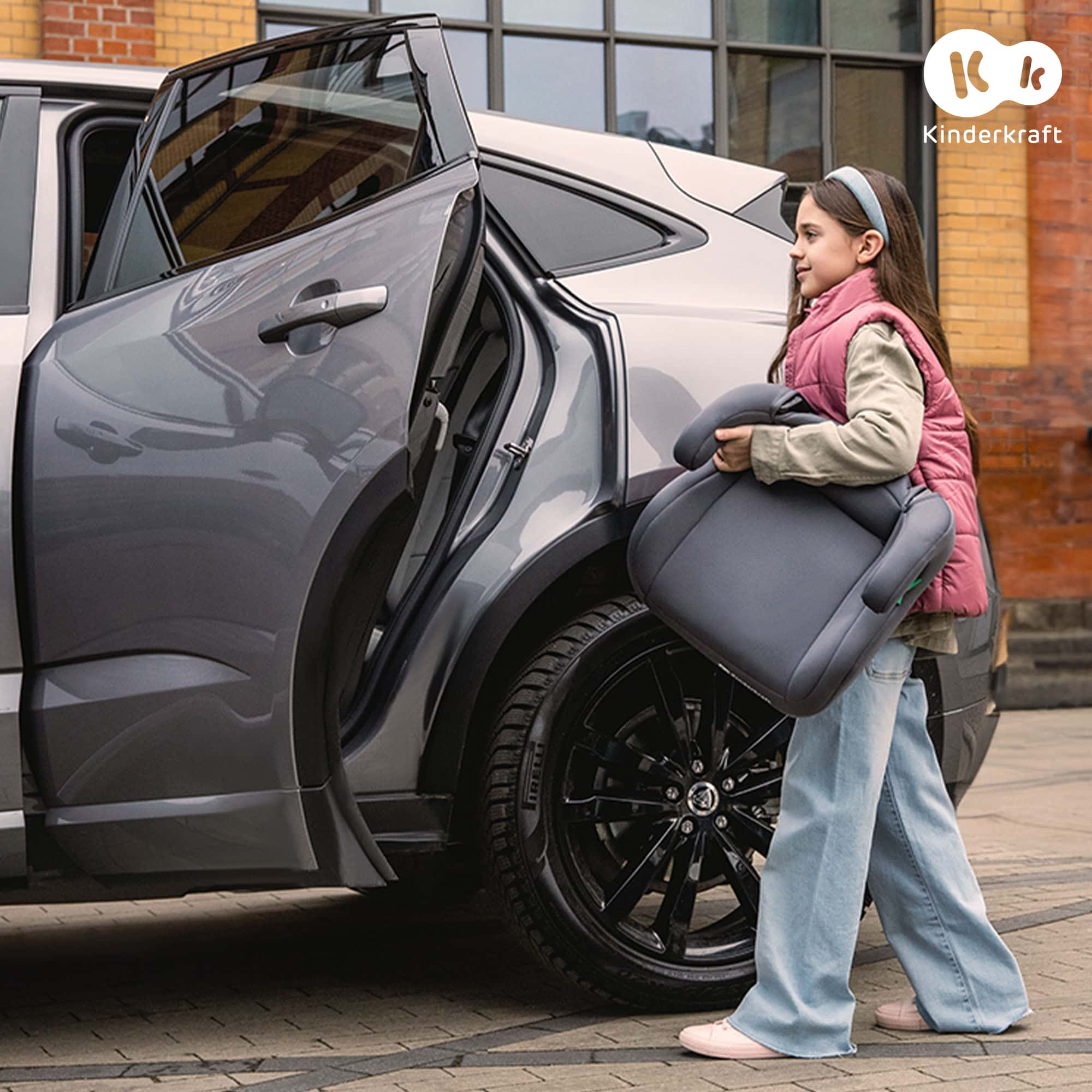 Ein Mädchen mit rosa Weste trägt einen Autositz neben einem grauen Auto.