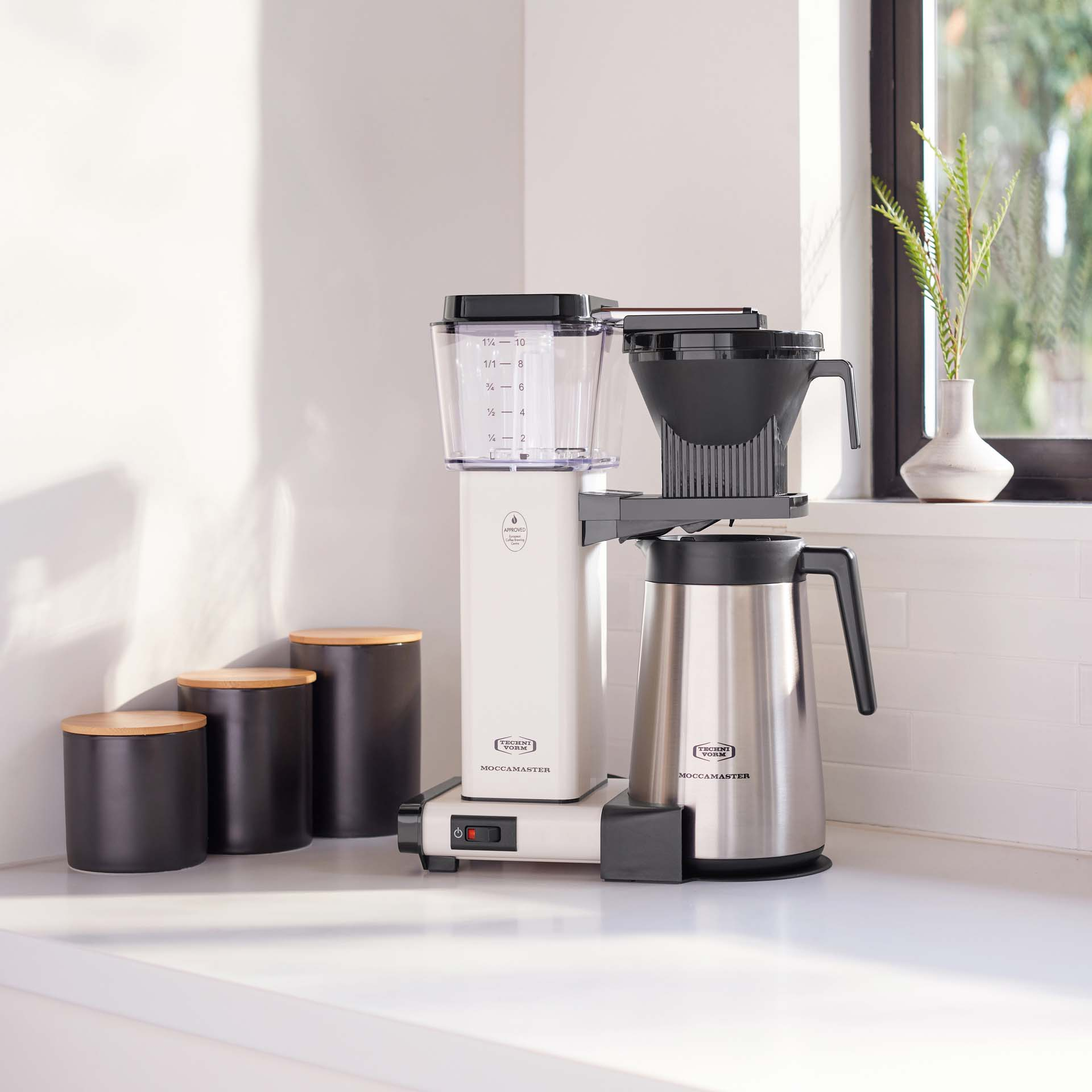 Una caffettiera bianca con caraffa argentata accanto a barattoli neri.