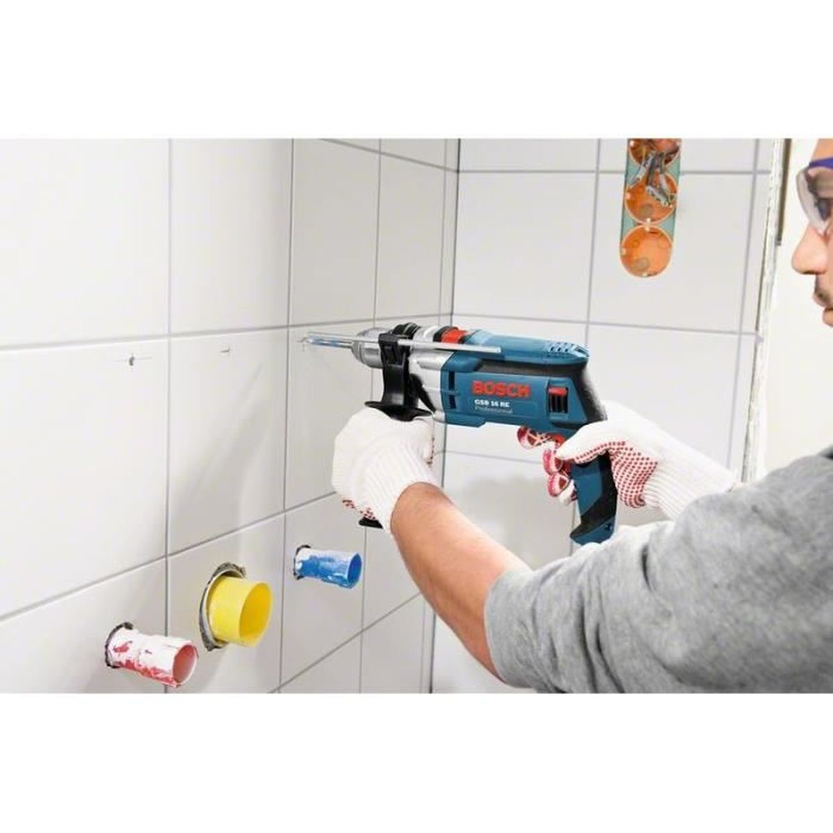 Mann bohrt mit Bosch-Bohrmaschine in weiße Fliesenwand, Schutzhandschuhe.