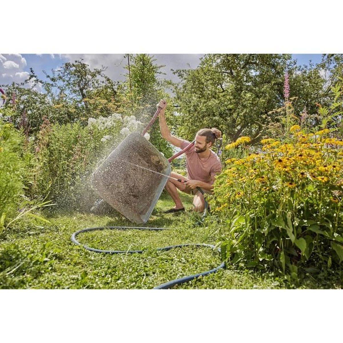 Mann reinigt eine Schubkarre mit einem Schlauch im Garten, umgeben von Pflanzen.