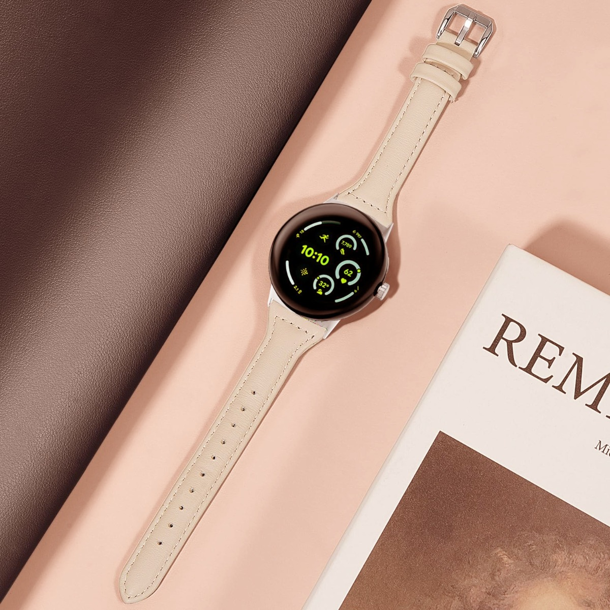Eine helle Smartwatch mit einem beigen Armband liegt auf einem rosafarbenen Untergrund.