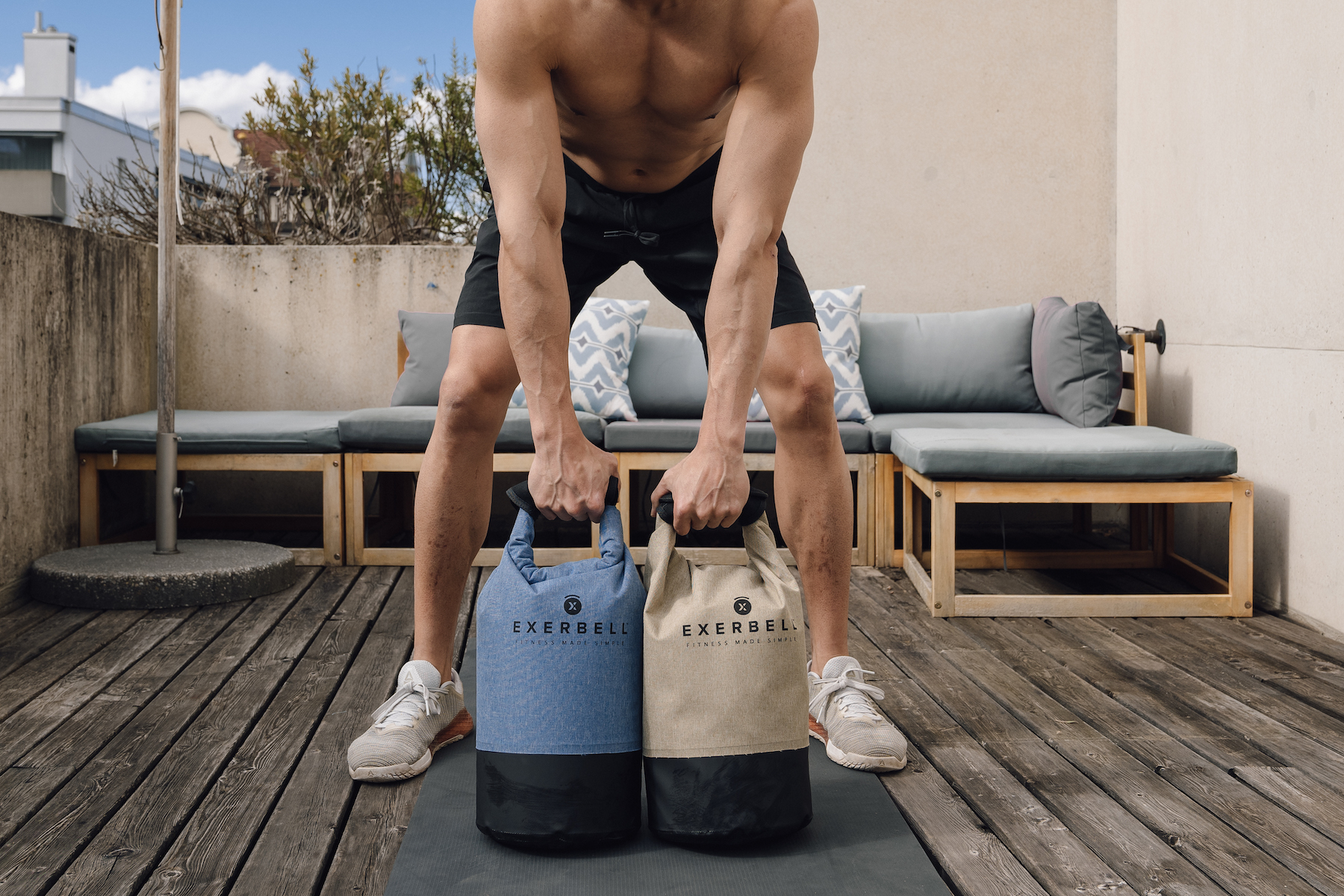 Un homme en short noir soulève deux sacs de poids, un bleu et un beige.