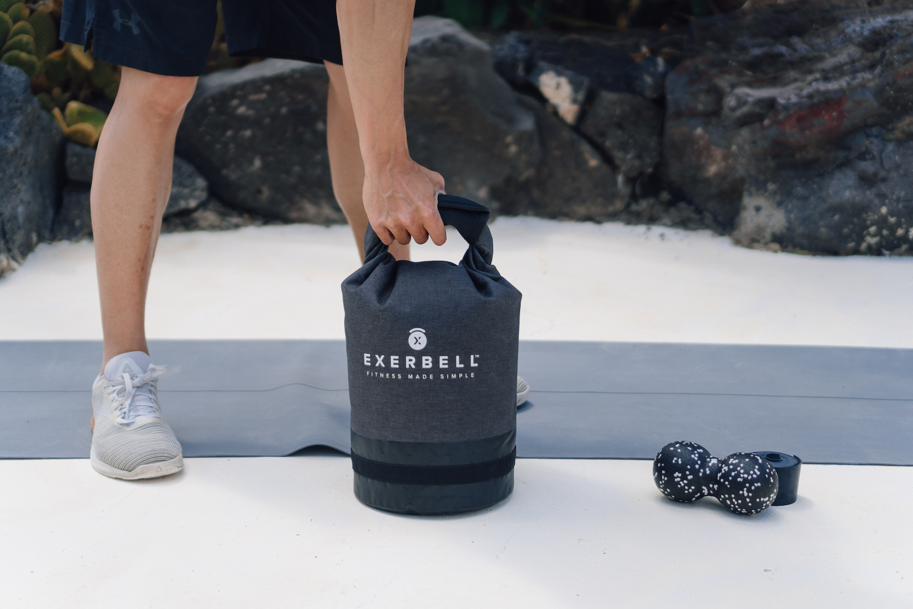 Un homme soulève une kettlebell grise avec logo "EXERBELL" sur un tapis.