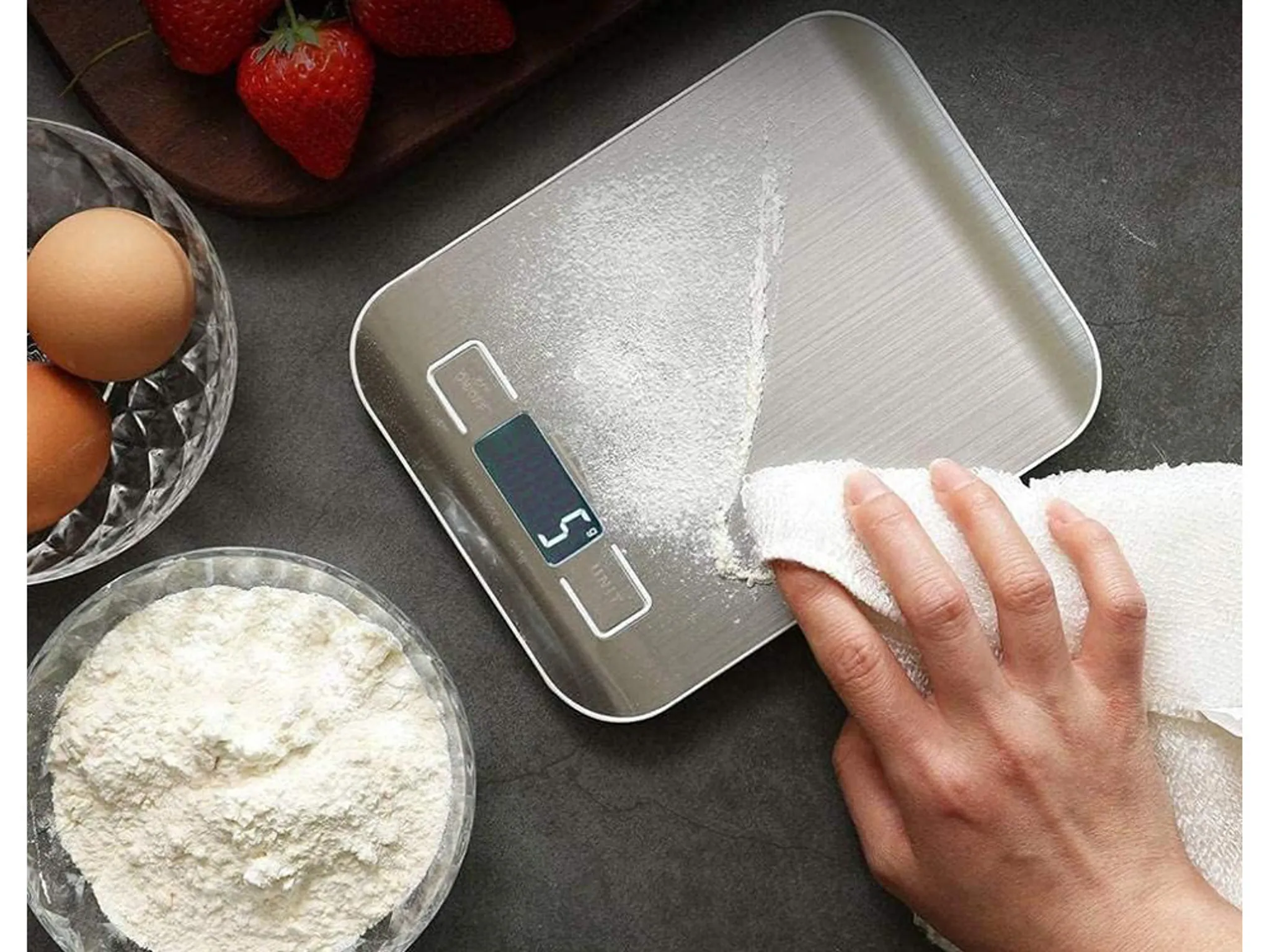 Eine Hand wischt Mehl von einer digitalen Küchenwaage. Eier und Erdbeeren liegen daneben.