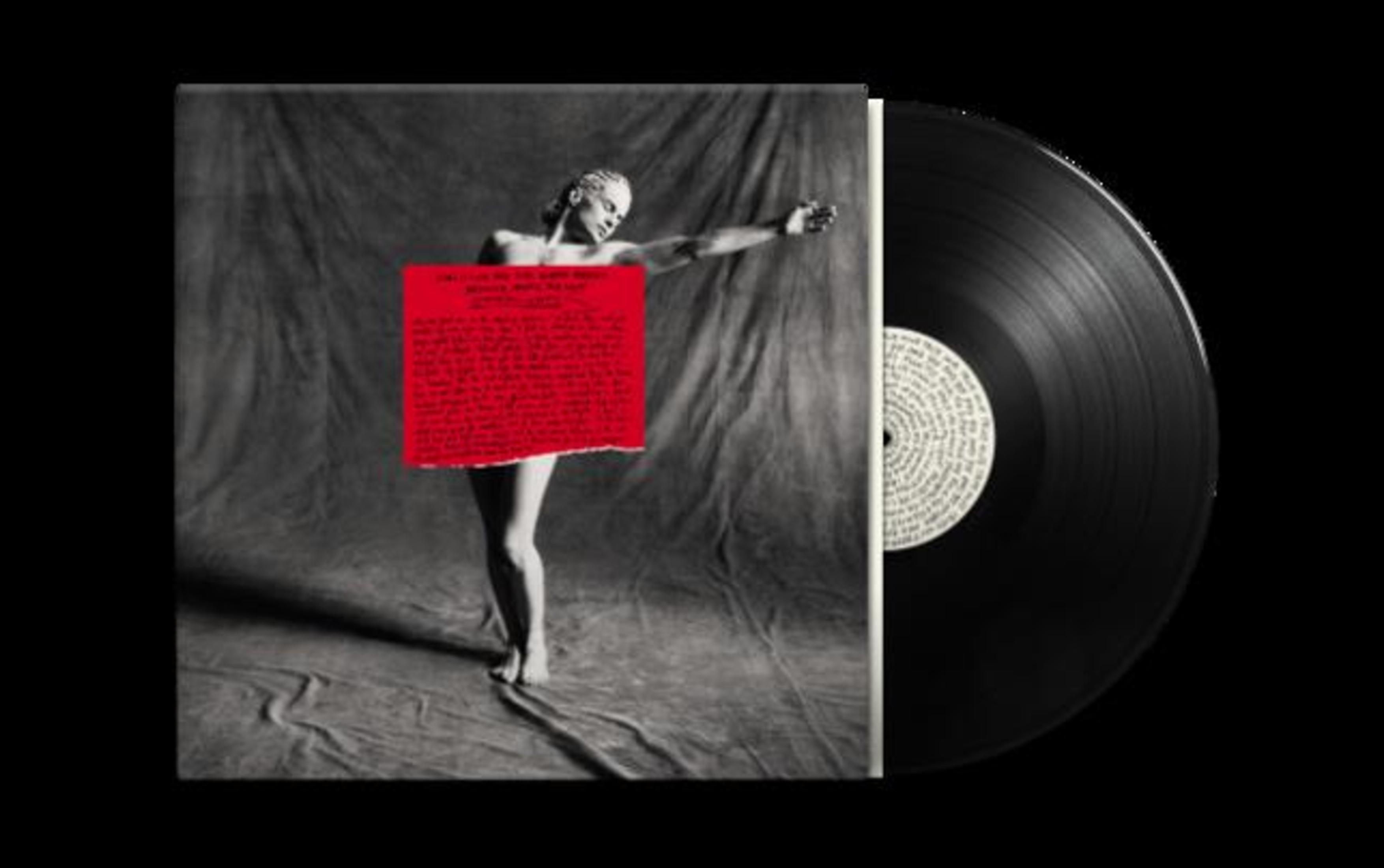 Una persona in piedi su uno sfondo grigio, tiene un cartello rosso con testo, accanto a un disco in vinile.