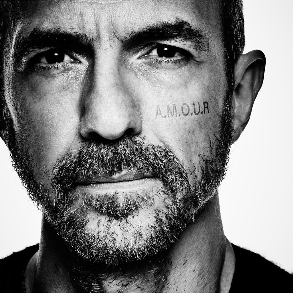Primo piano di un uomo con la barba, la parola AMOUR tatuata sulla guancia.