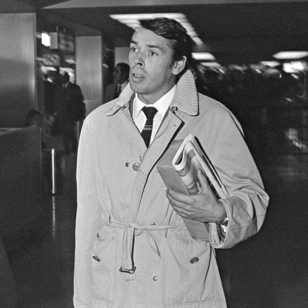 Een man in een trenchcoat en stropdas houdt boeken en kranten vast.