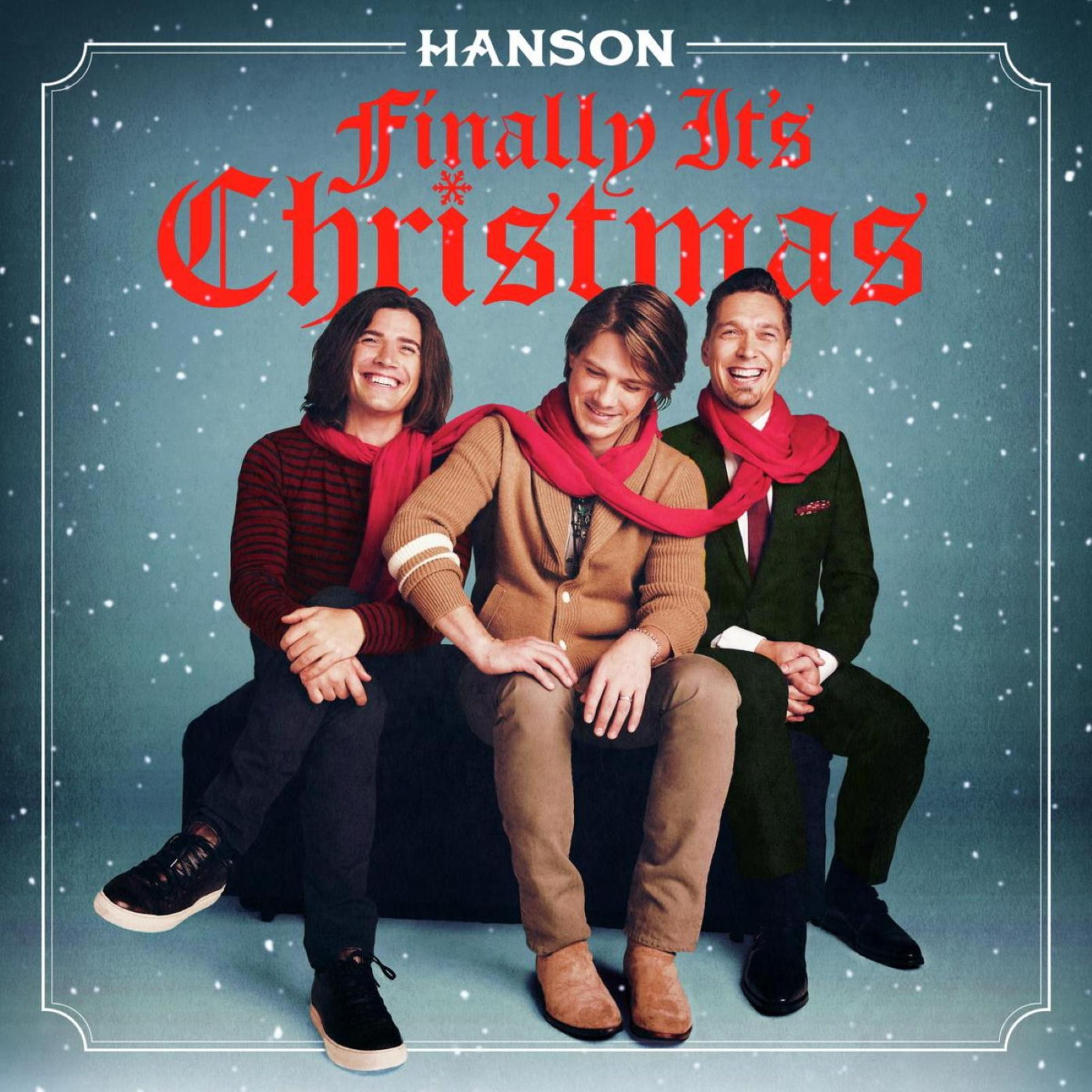 Tre uomini in abiti festivi sorridono su uno sfondo blu e nevoso. Il testo dice 'Hanson Finally It's Christmas'.