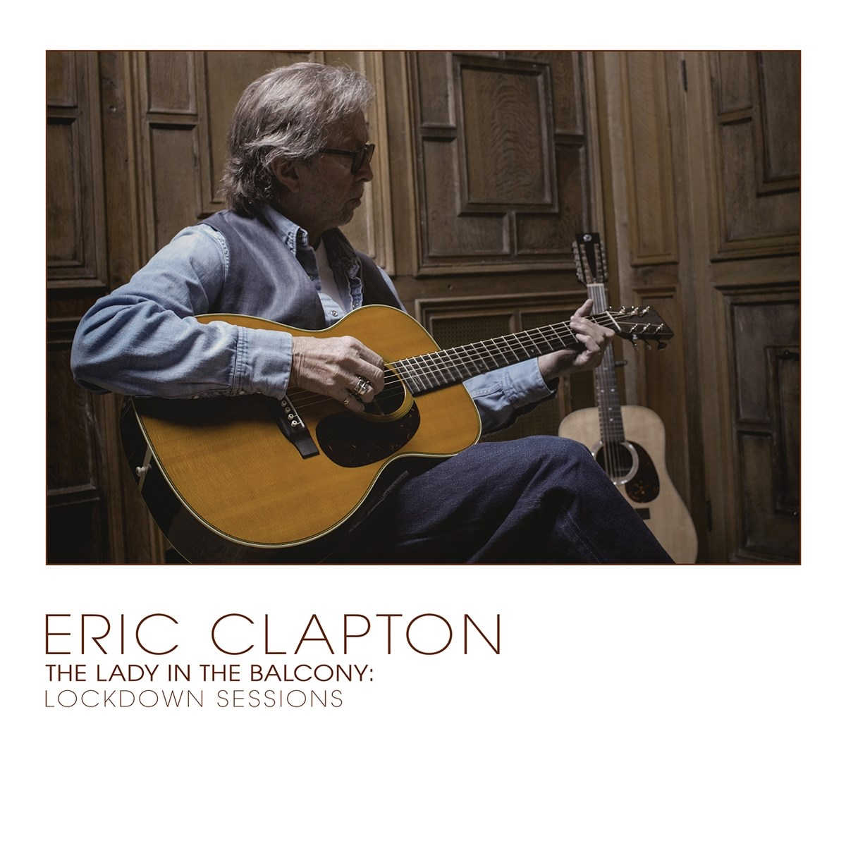 Eric Clapton suona la chitarra acustica contro un muro di legno, un'altra chitarra sullo sfondo.