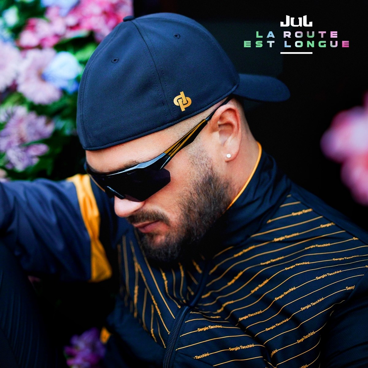 Uomo con cappellino e occhiali da sole indossa giacca sportiva a righe. Fiori dietro.