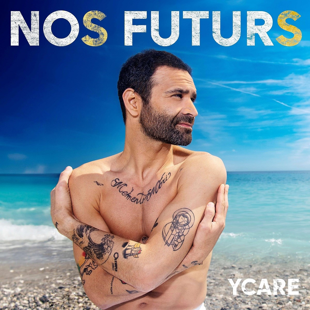 Uomo con tatuaggi si abbraccia su una spiaggia vicino al mare.