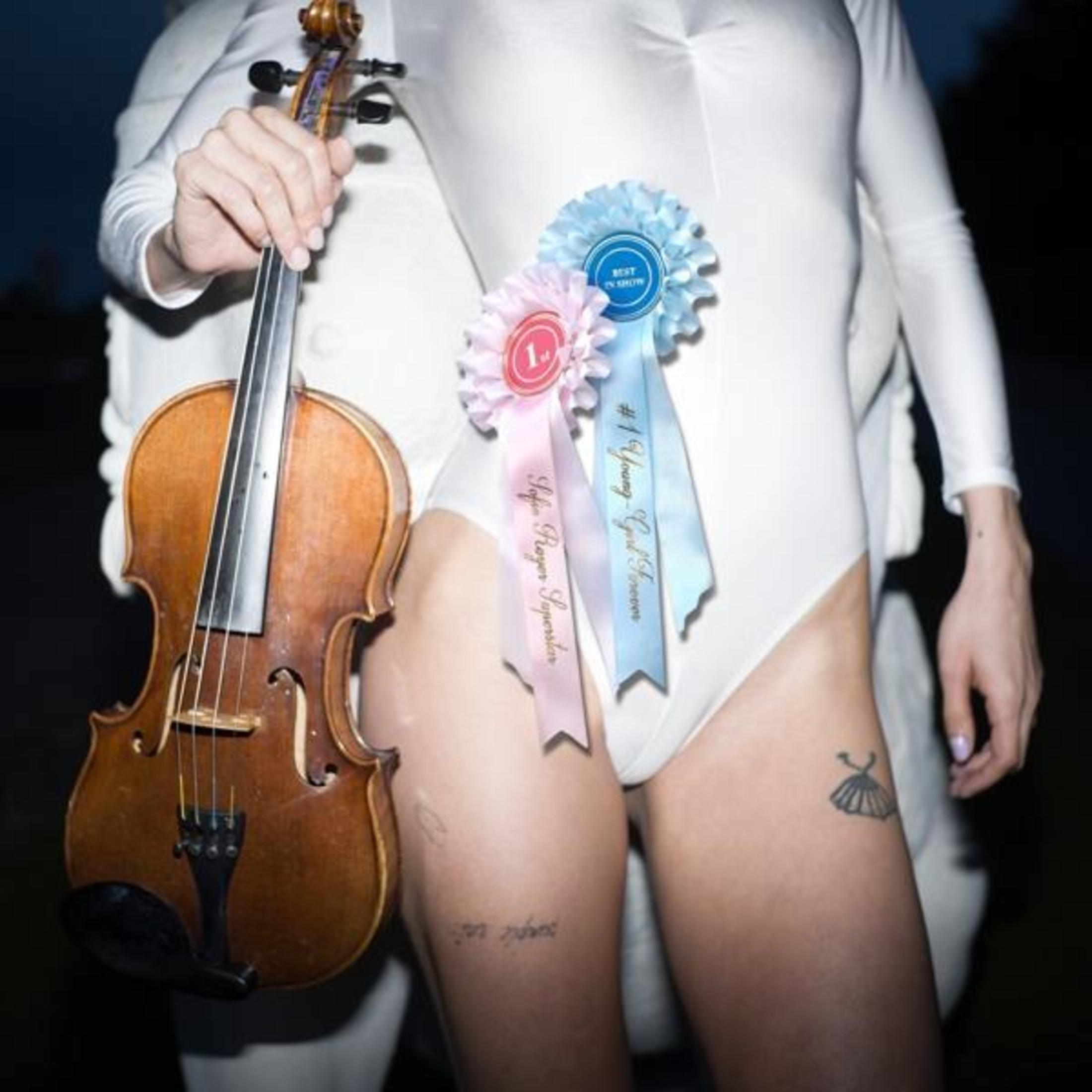 Una persona in bianco tiene un violino. Nastri dicono '1' e 'Migliore in mostra'.