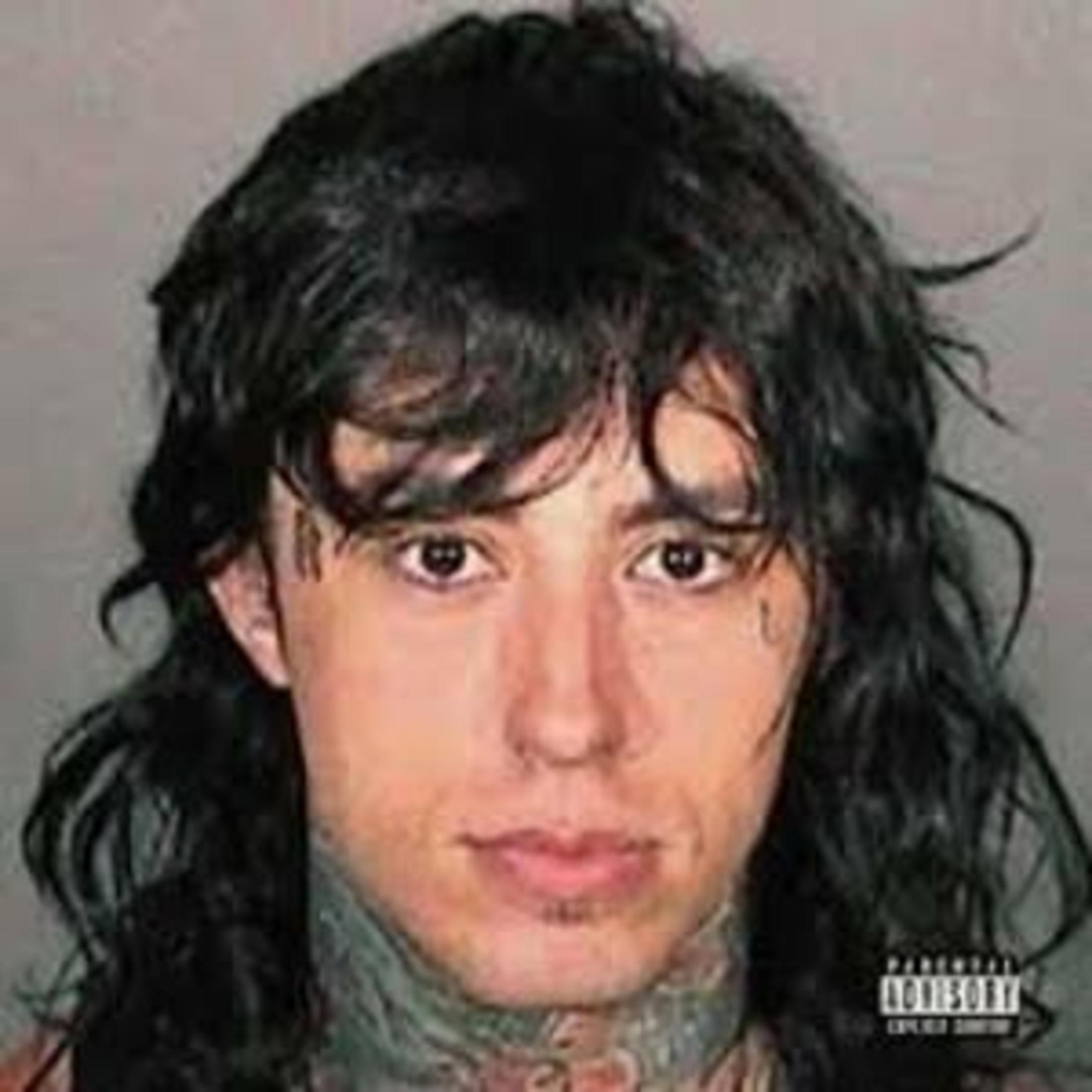 Un uomo con capelli scuri fino alle spalle guarda avanti. Ha tatuaggi sul collo.