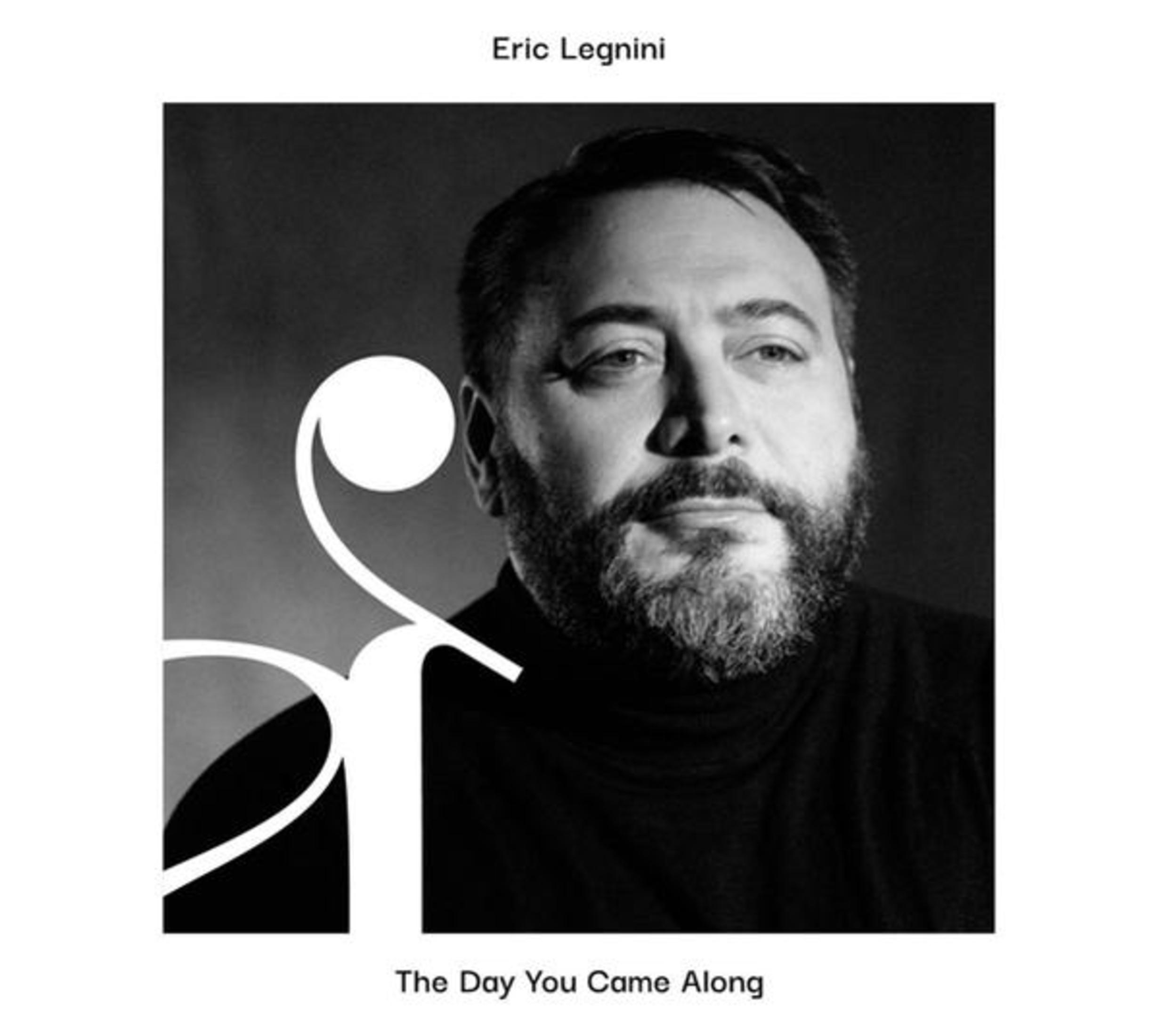Un uomo con la barba indossa un dolcevita nero. Un grande simbolo bianco è a sinistra.