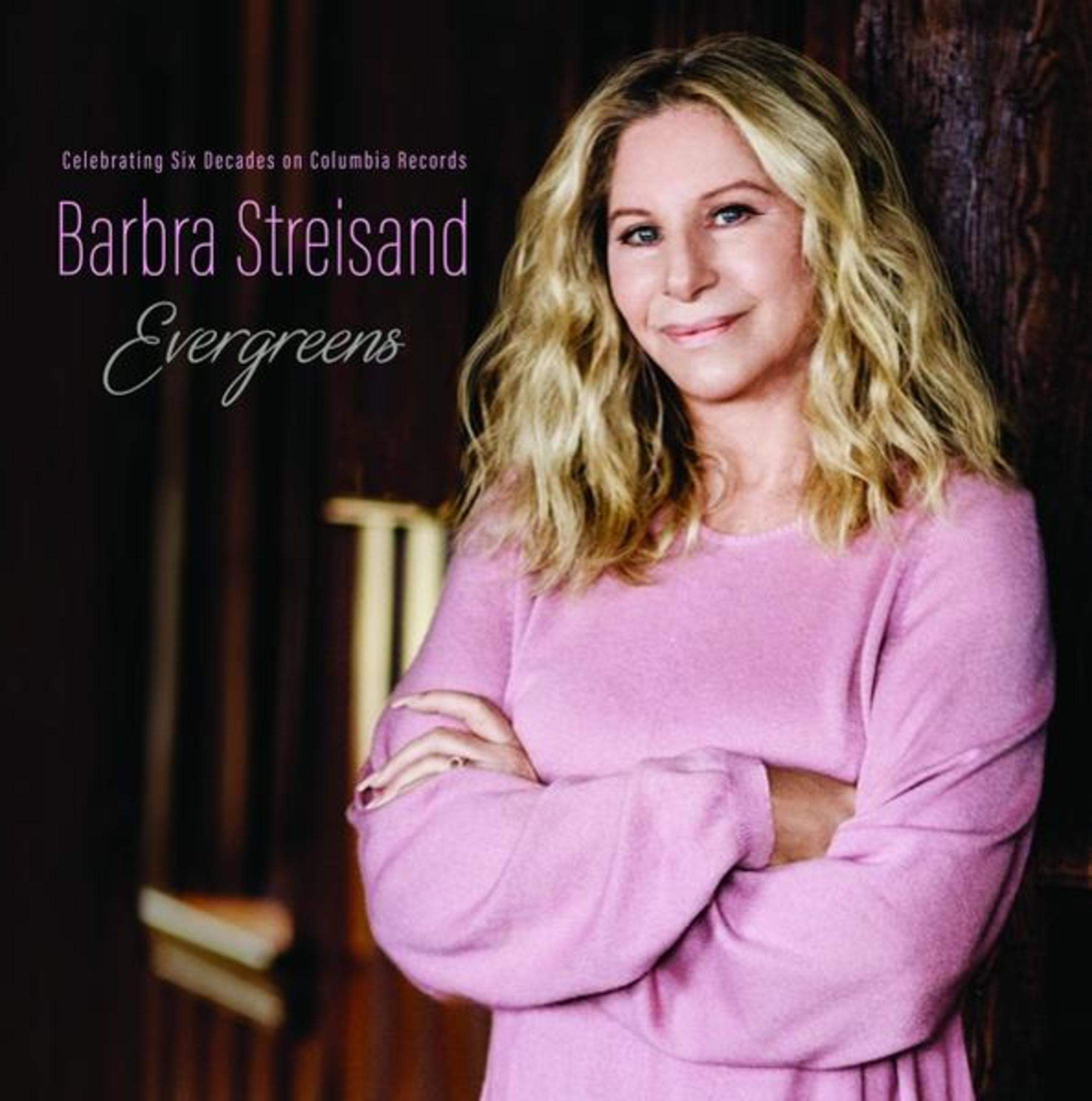 Barbra Streisand in maglione rosa, braccia incrociate, su sfondo scuro.