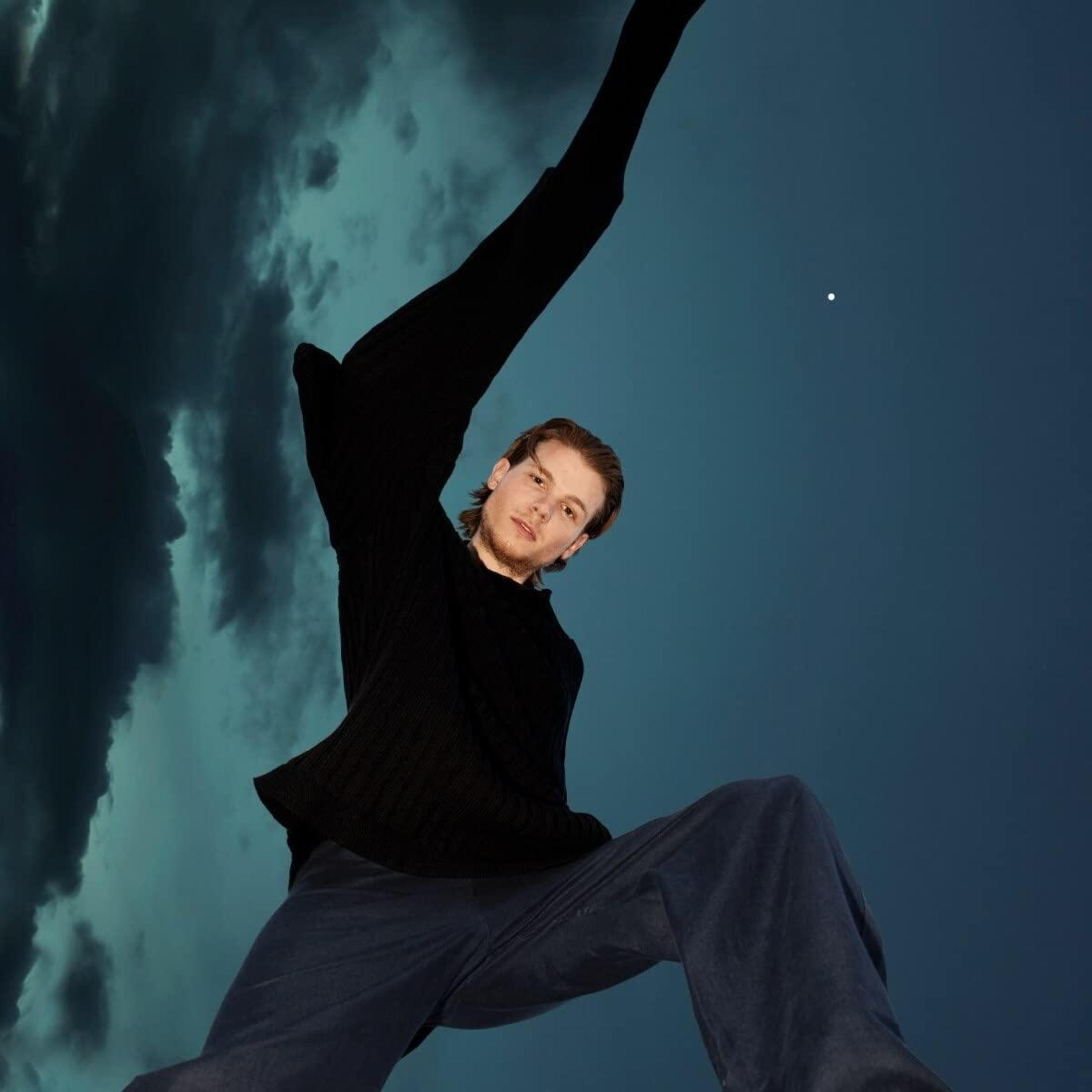 Un uomo con maglione nero e pantaloni blu si erge contro un cielo scuro.