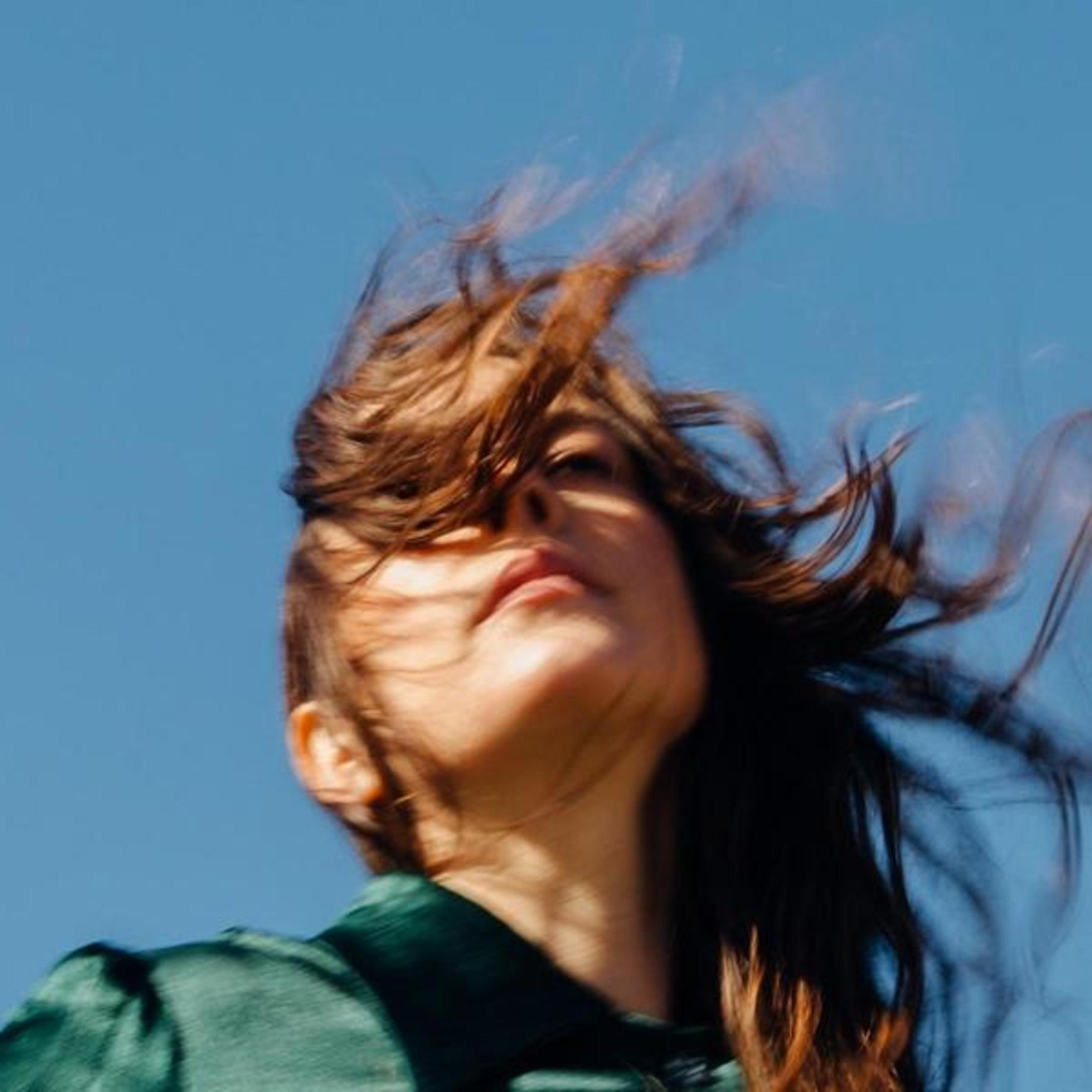Donna con capelli castani al vento su cielo blu, indossa abiti verdi.