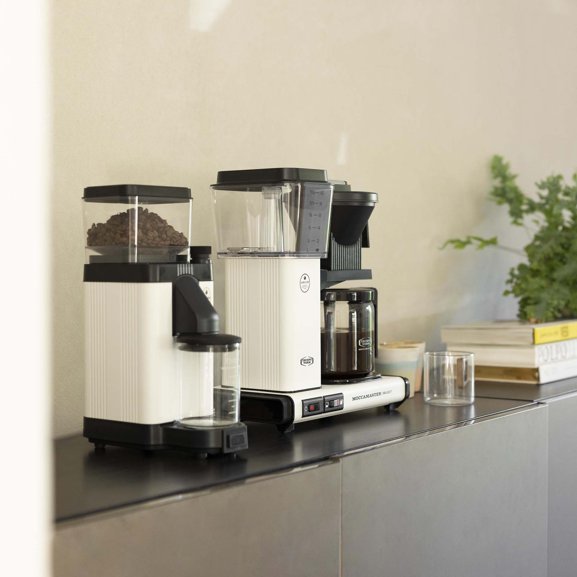 Zwei weiße Kaffeemaschinen und eine Kaffeemühle mit Kaffeebohnen auf dunkler Oberfläche.