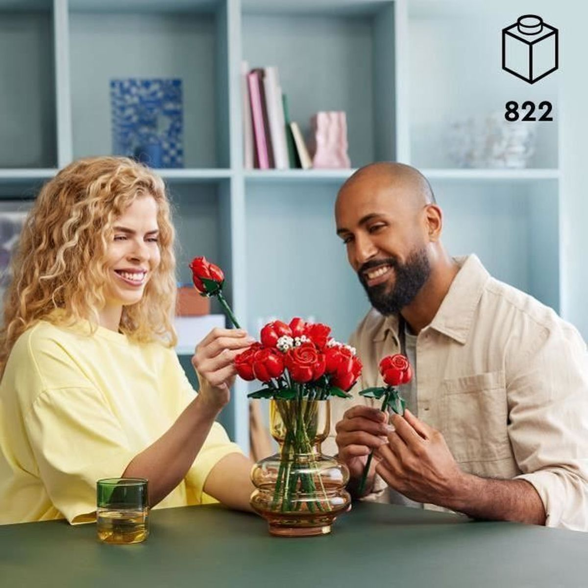 Een stel bouwt rode LEGO rozen in een glazen vaas op een tafel.