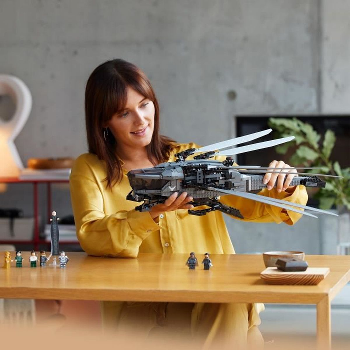 Kobieta trzyma szary statek kosmiczny Lego. Kilka małych figurek stoi na stole.