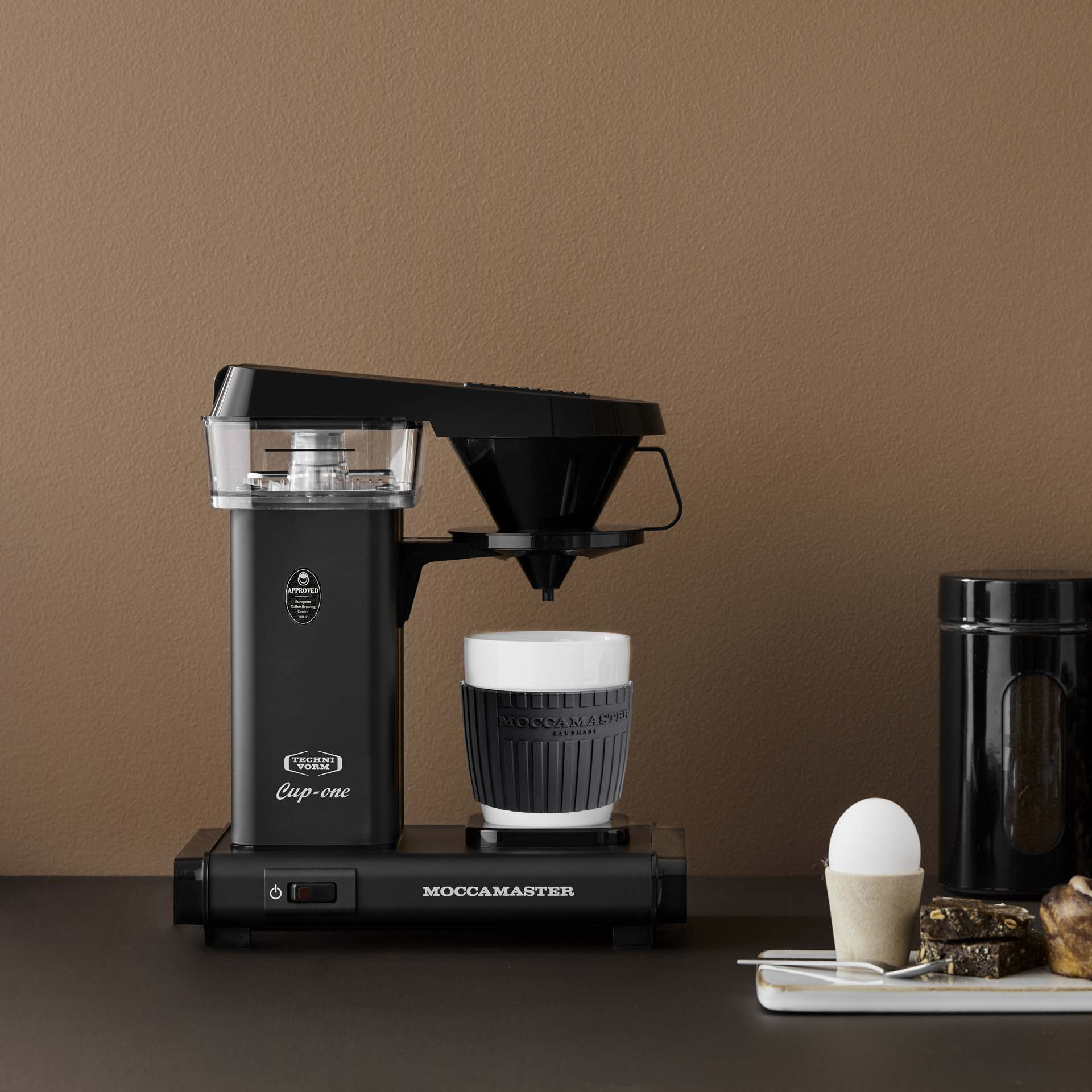 Black coffee maker with cup, next to it an egg in a holder and brownies.