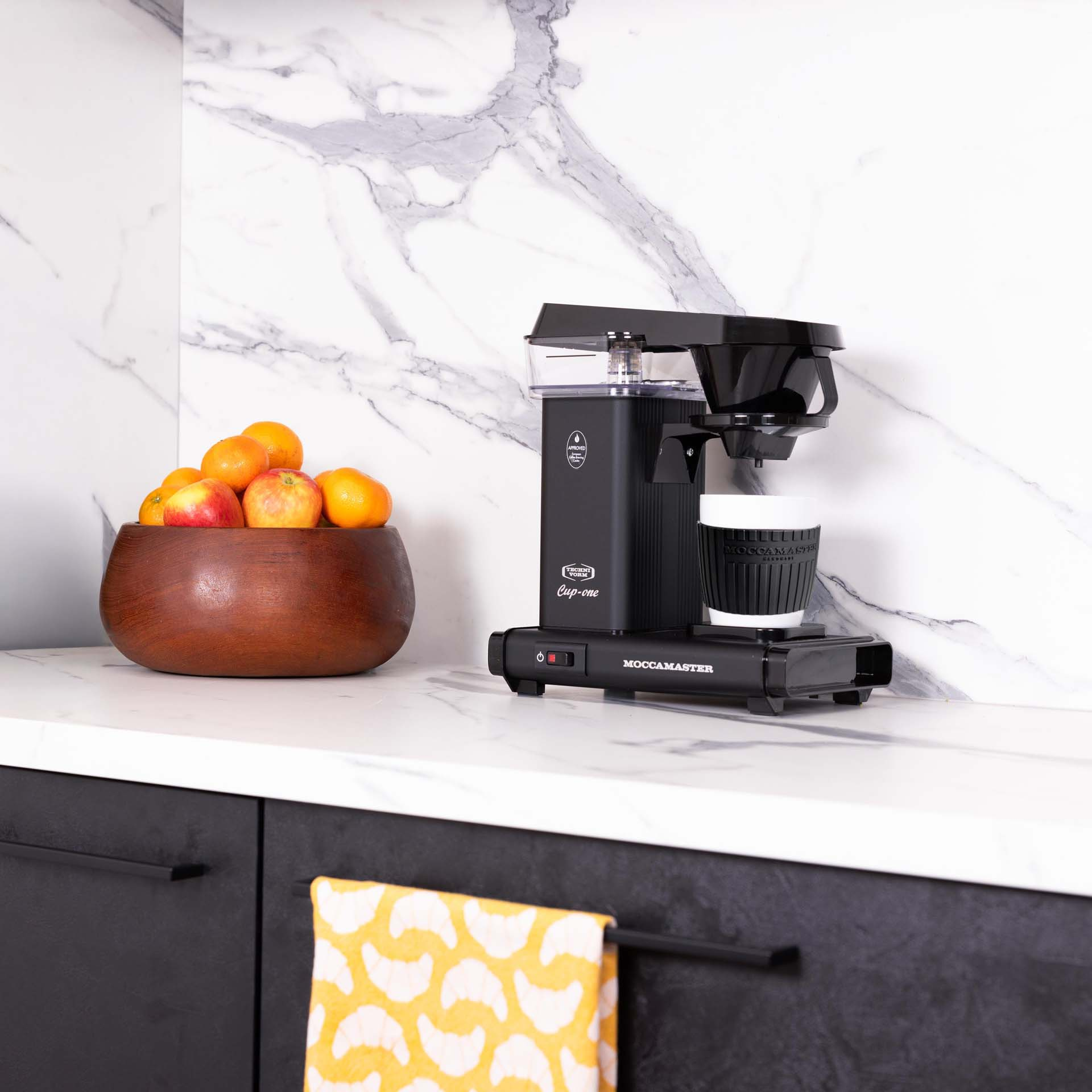 Black coffee maker next to a wooden bowl filled with apples and oranges.